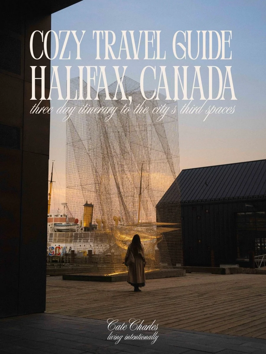 text reads Cozy Travel Guide Halifax, Canada three day itinerary to the city's third spaces, living intentionally. The image is of a girl wearing a long trench coat standing on a waterfront boardwalk looking at a modern art installation at blue hour