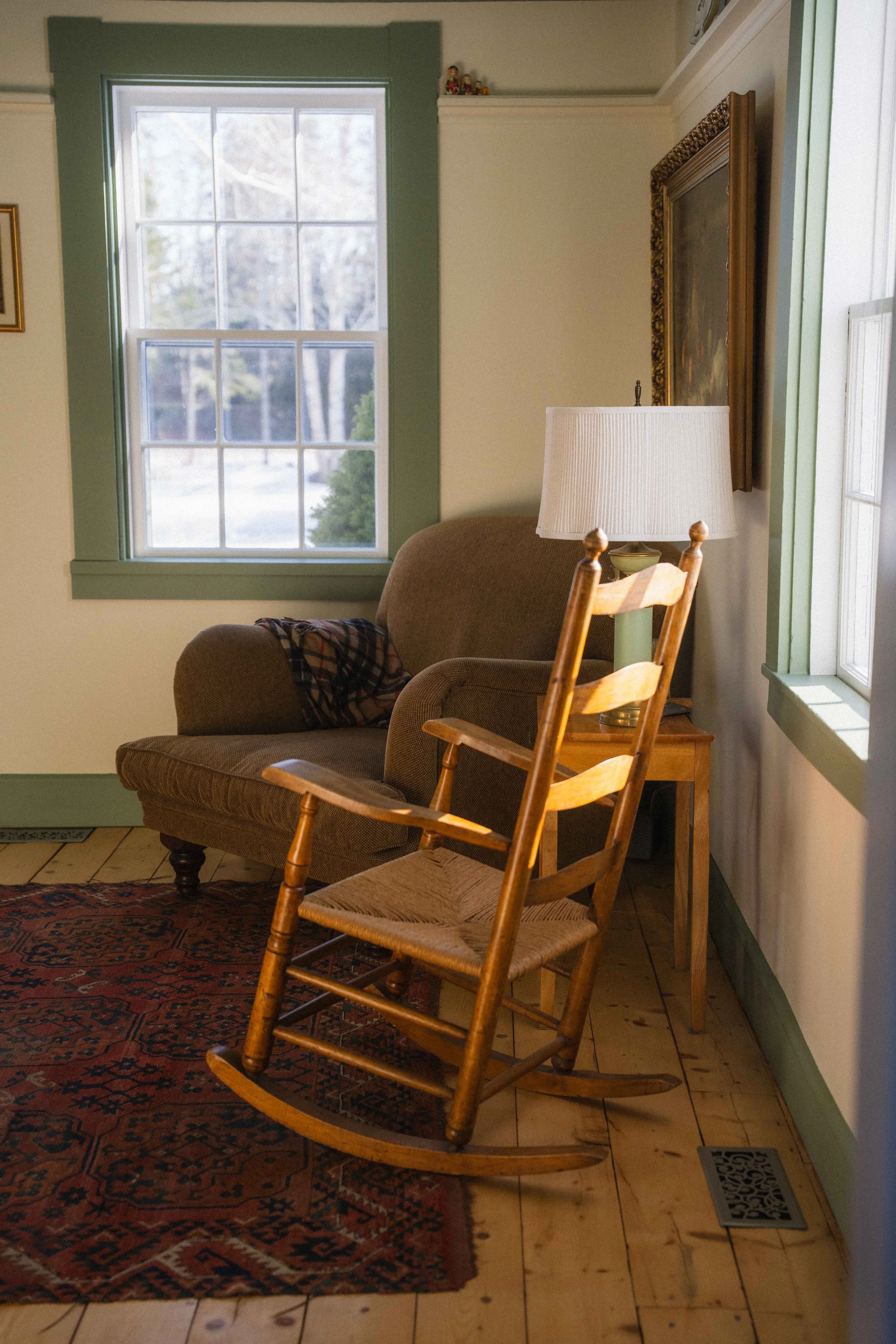 cozy cottage living room with ladder back rocking chair with wicker seat and roll arm english style armchair and woven wool turkish rug and antique oil painting in a cream room with green painted trim.