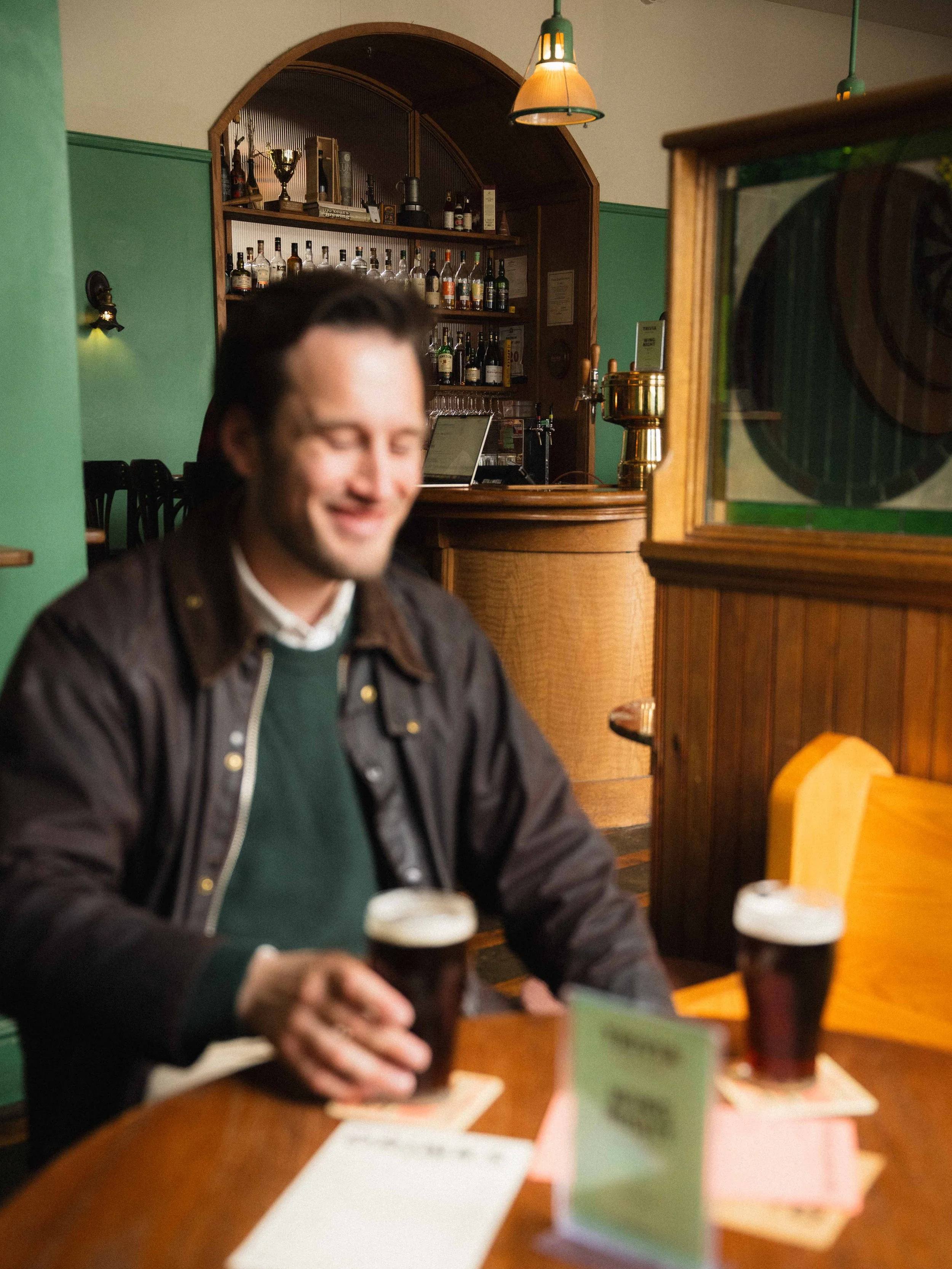 a man sitting in a local pub in Halifax Nova Scotia holding a pint of beer and smiling, he is wearing a barbour jacket with sweater and is blurred out, while the bar with cask ale is in focus