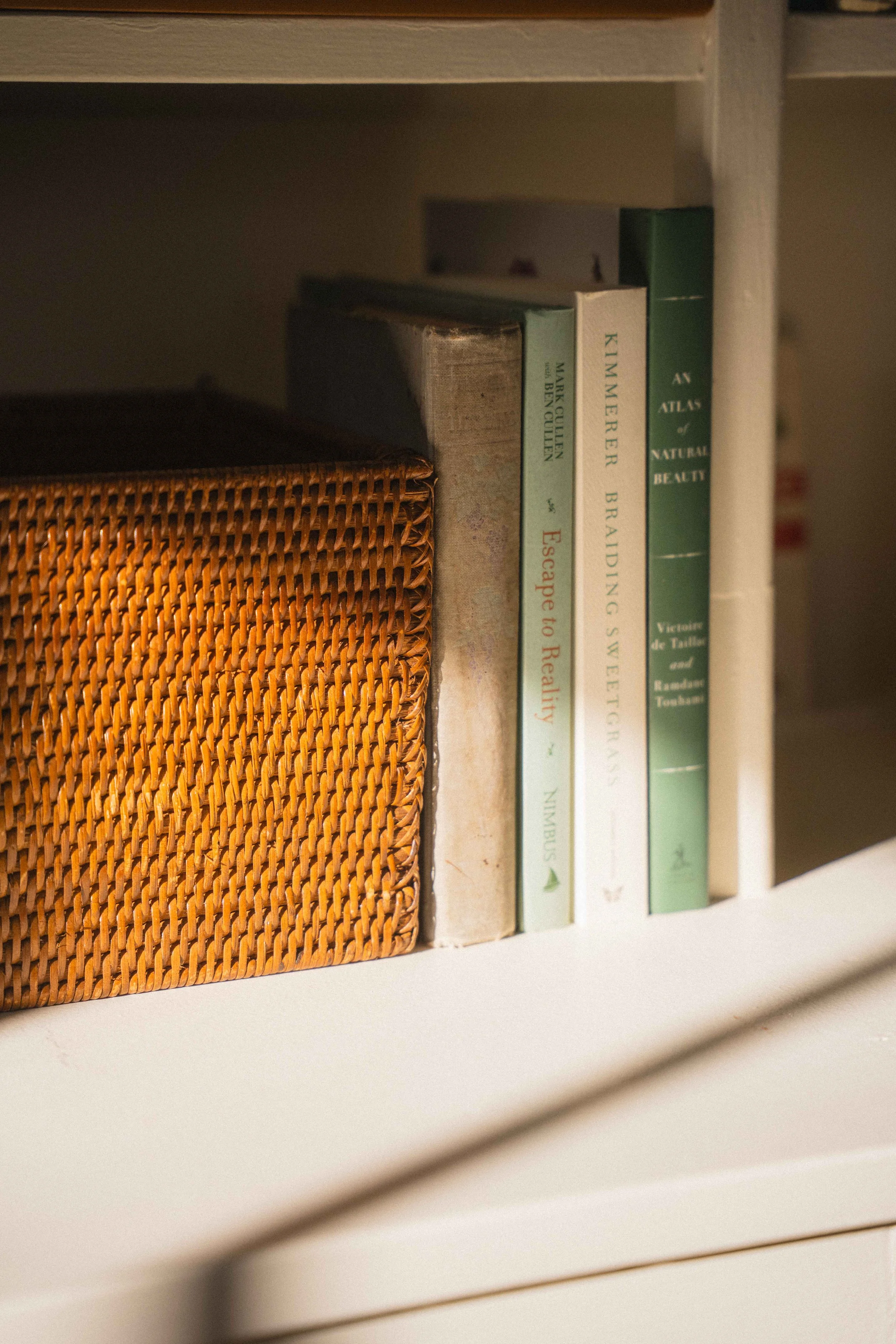 sunlight on a cream bookshelf with books braiding sweetgrass, an atlas of natural beauty, escape to reality gardening book, and a wicker basket for a cottagecore library