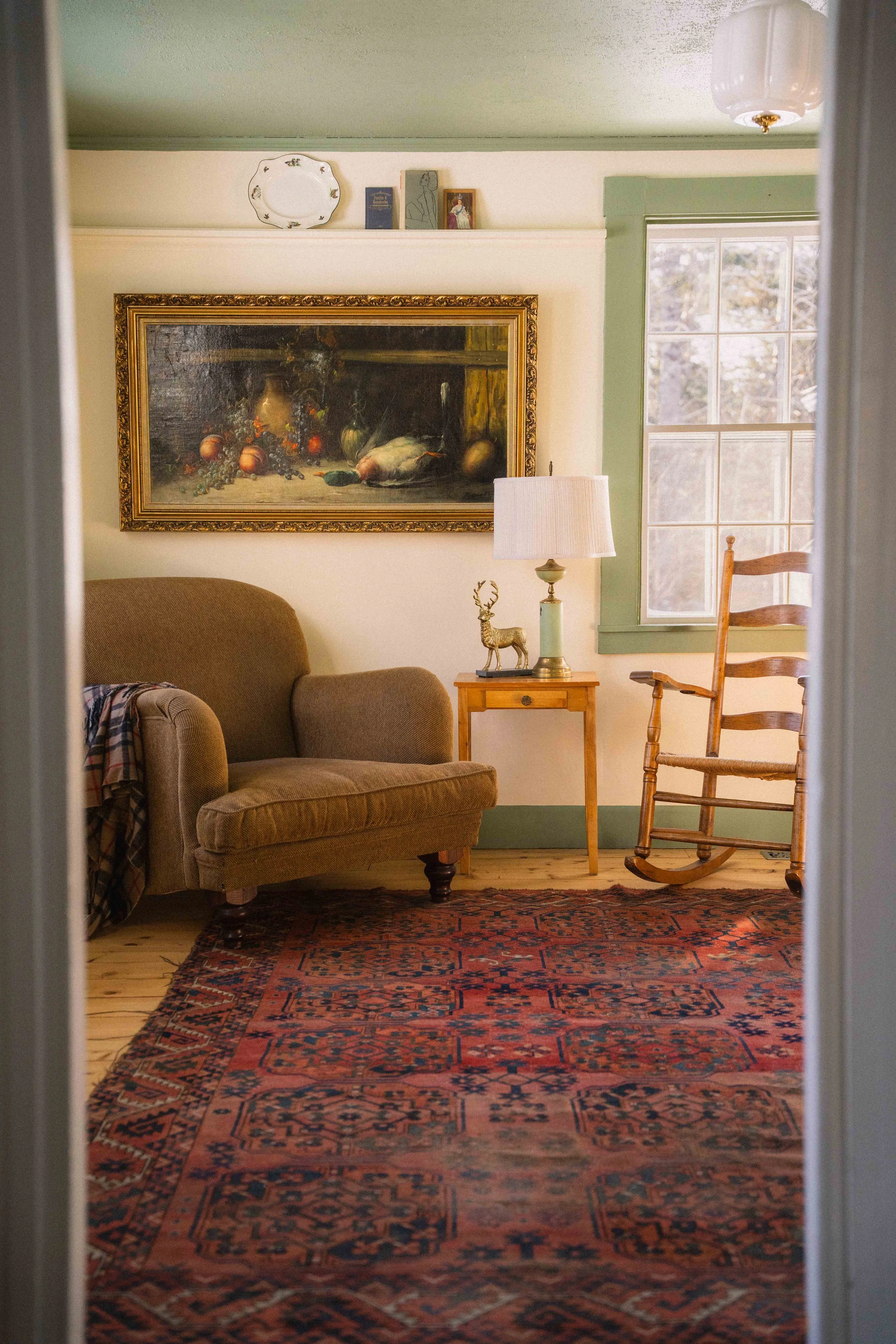 cottagecore interior with antique wool turkish rug, ladder back rocking chair, scalloped lamp, still life oil painting, english roll arm chair, and tartan throw blanket, historic home with crown molding and green trim with cream walls