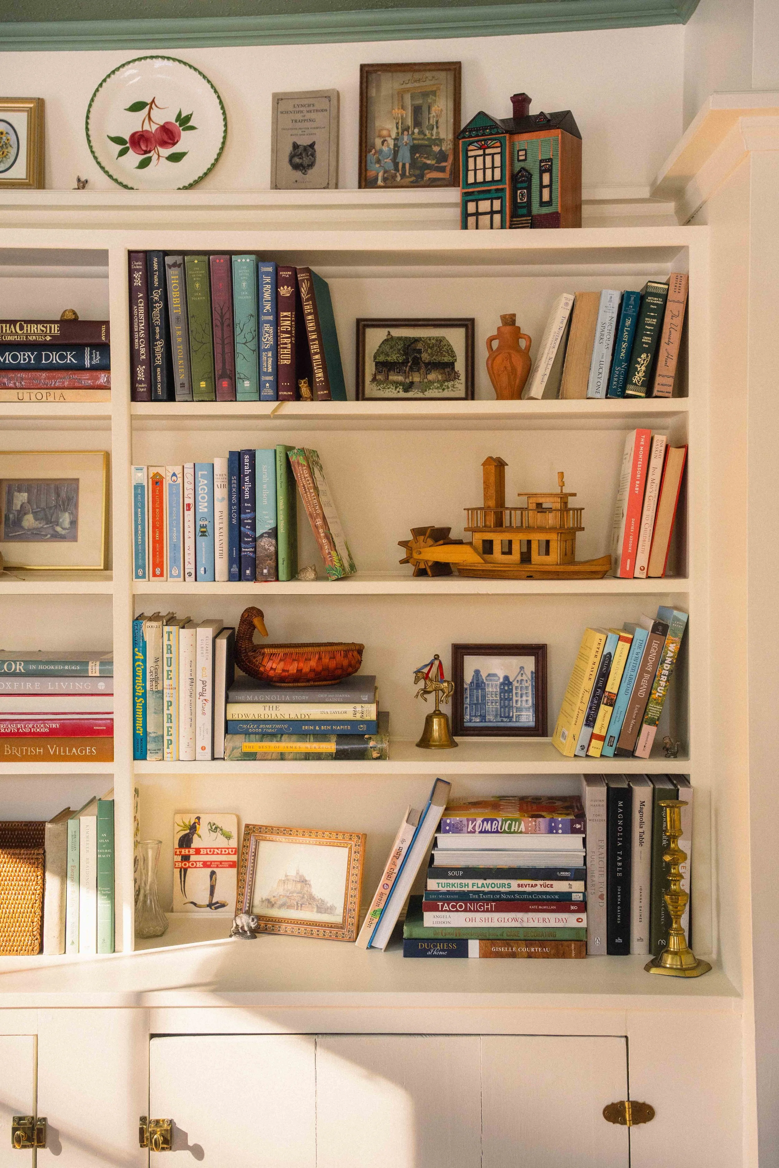 colourful cottage library built-in bookshelf with cream and green paint, collected antique finds. A wooden boat, a delft tile, a duck basket, a brass bell and candle stick, a ceramic house.