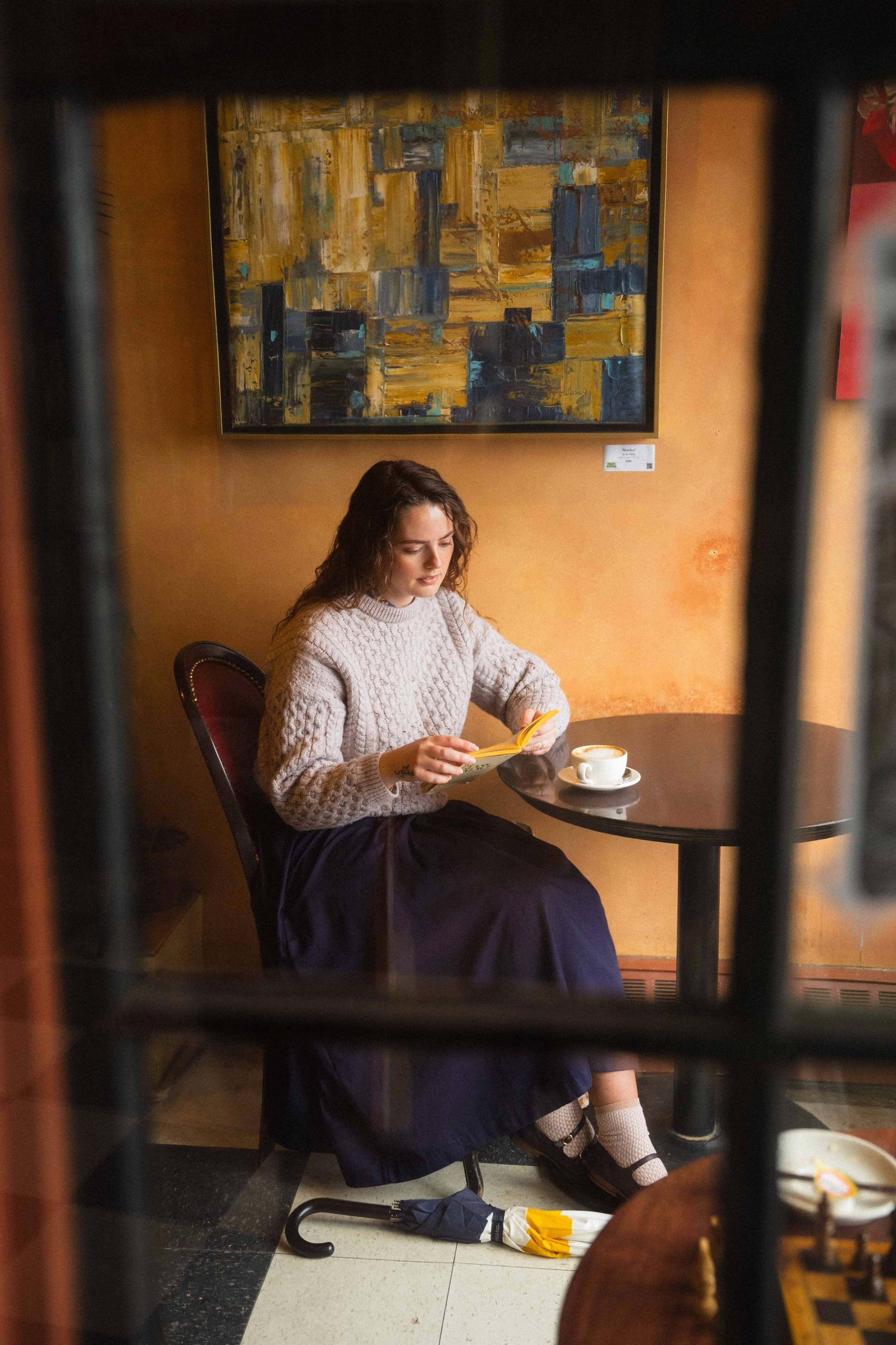 a view through a window of a girl sitting in a cafe, reading an old paperback book with a cappuccino and wearing a vintage irish wool sweater with a pleated skirt and mary jane shoes in a dark academia style