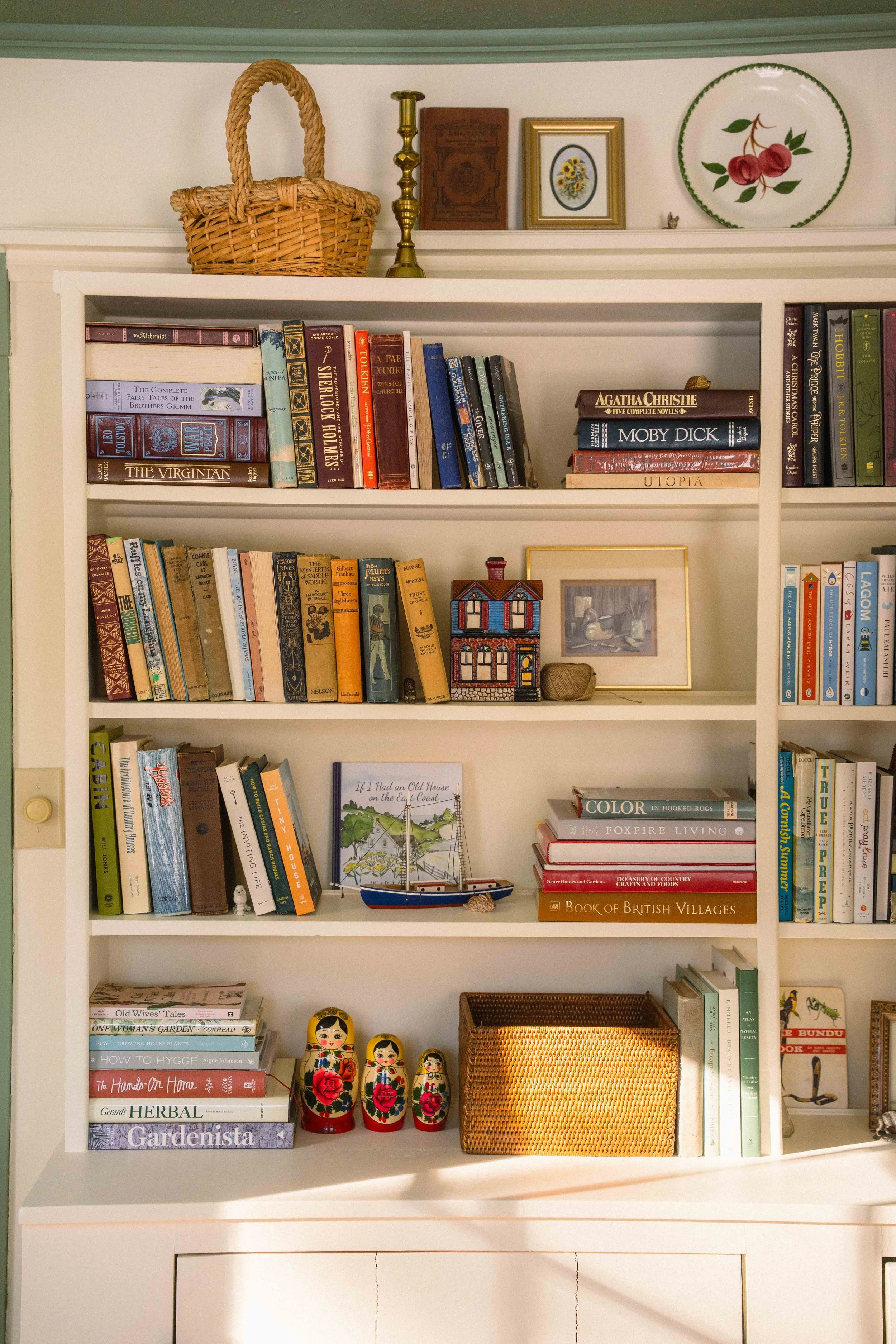 a styled bookshelf with colourful books and antique finds, including a woven basket, a brass candlestick, a ceramic house, matryoshka dolls, a miniature boat, and a book titles "if I had an old house on the east coast" and other home decor books