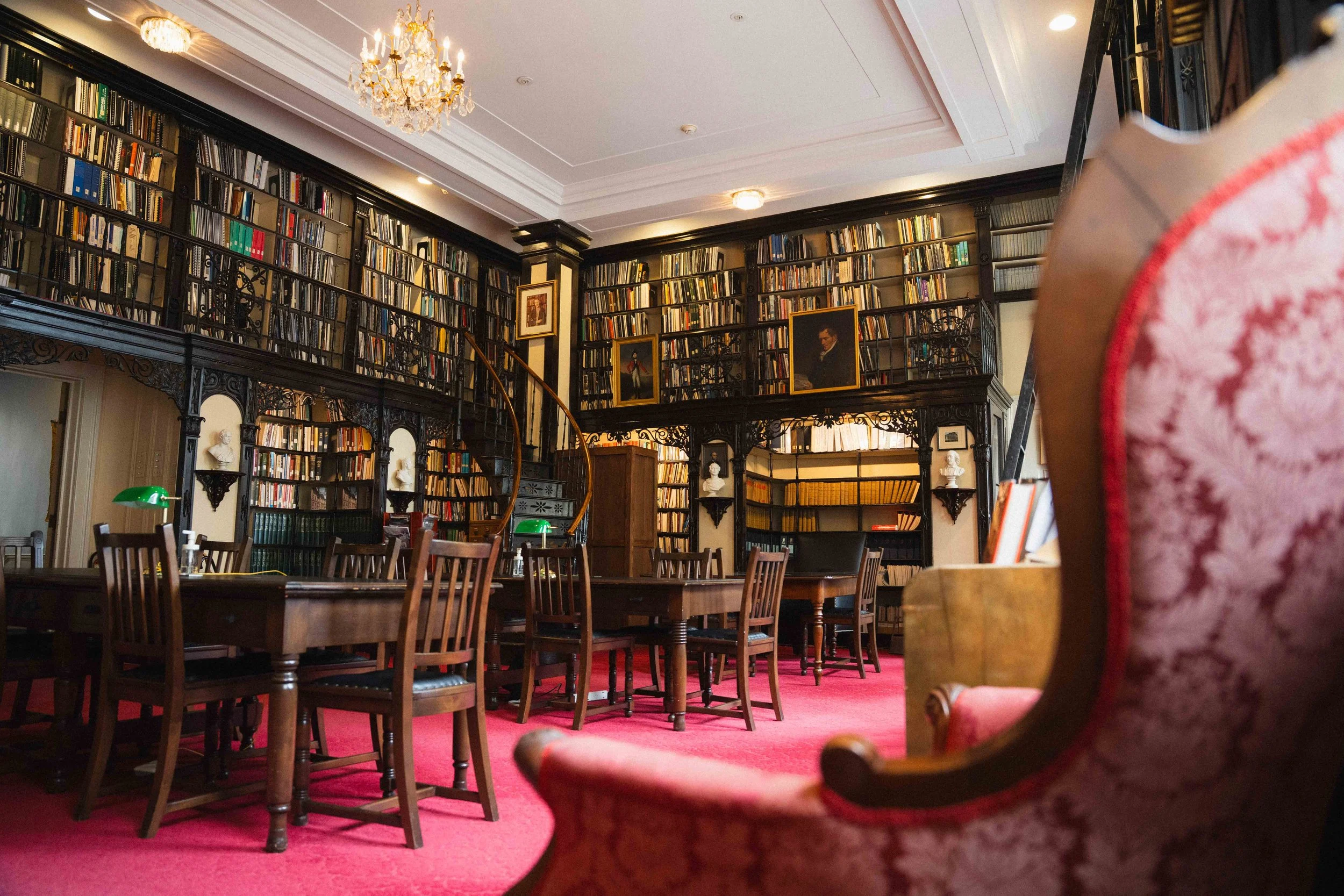a wide shot from behind an antique armchair of a historic library space in a provincial legislative building in Halifax, Canada with wooden study tables and a spiral staircase with walls lined with books and marble busts and historic portraits