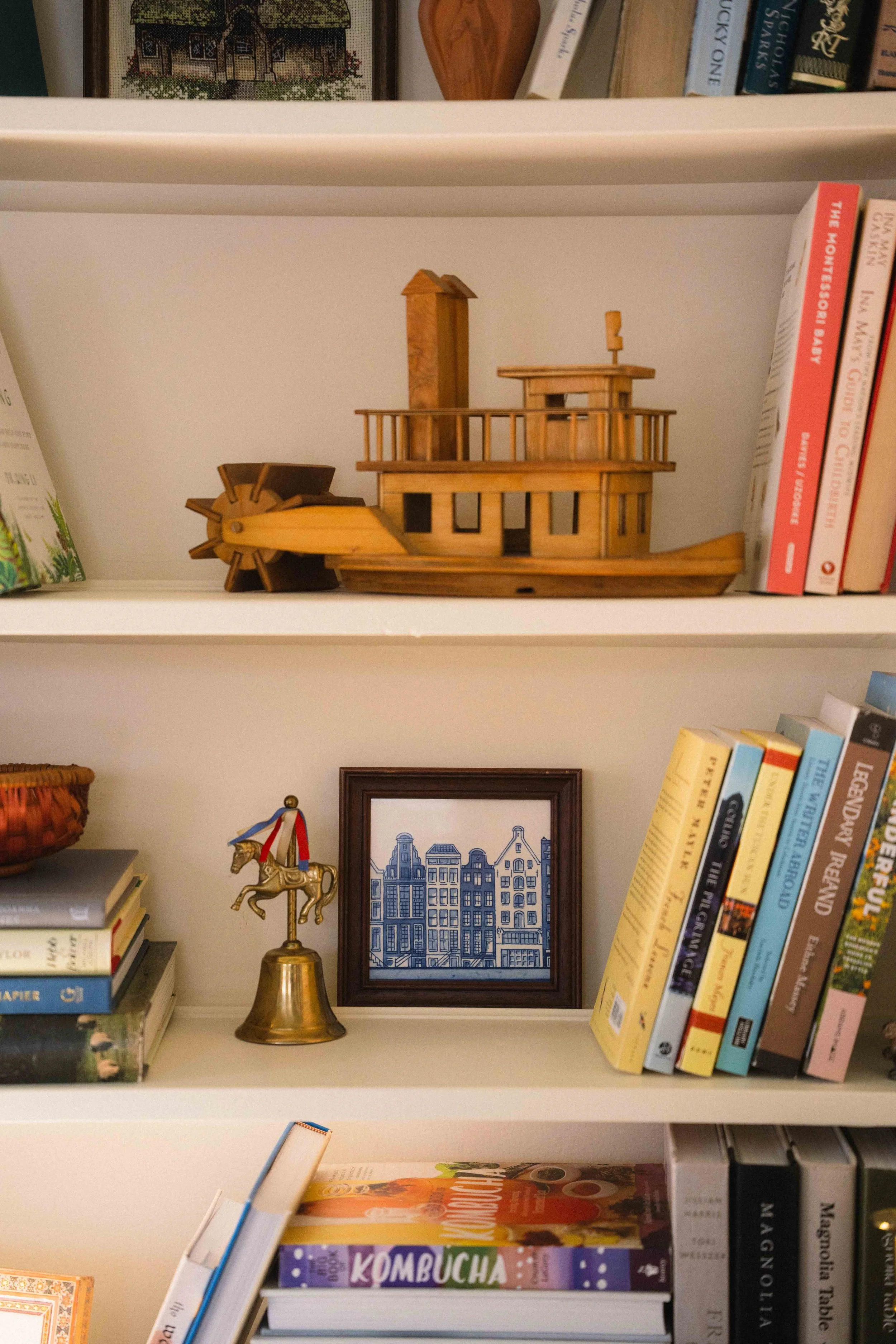 a cream bookshelf with antique collectibles featuring a framed blue and white delft tile, a brass bell, a wooden boat, and book titles including Under the Tuscan Sun, a Summer in Provence and a cross stitch of an cottage home