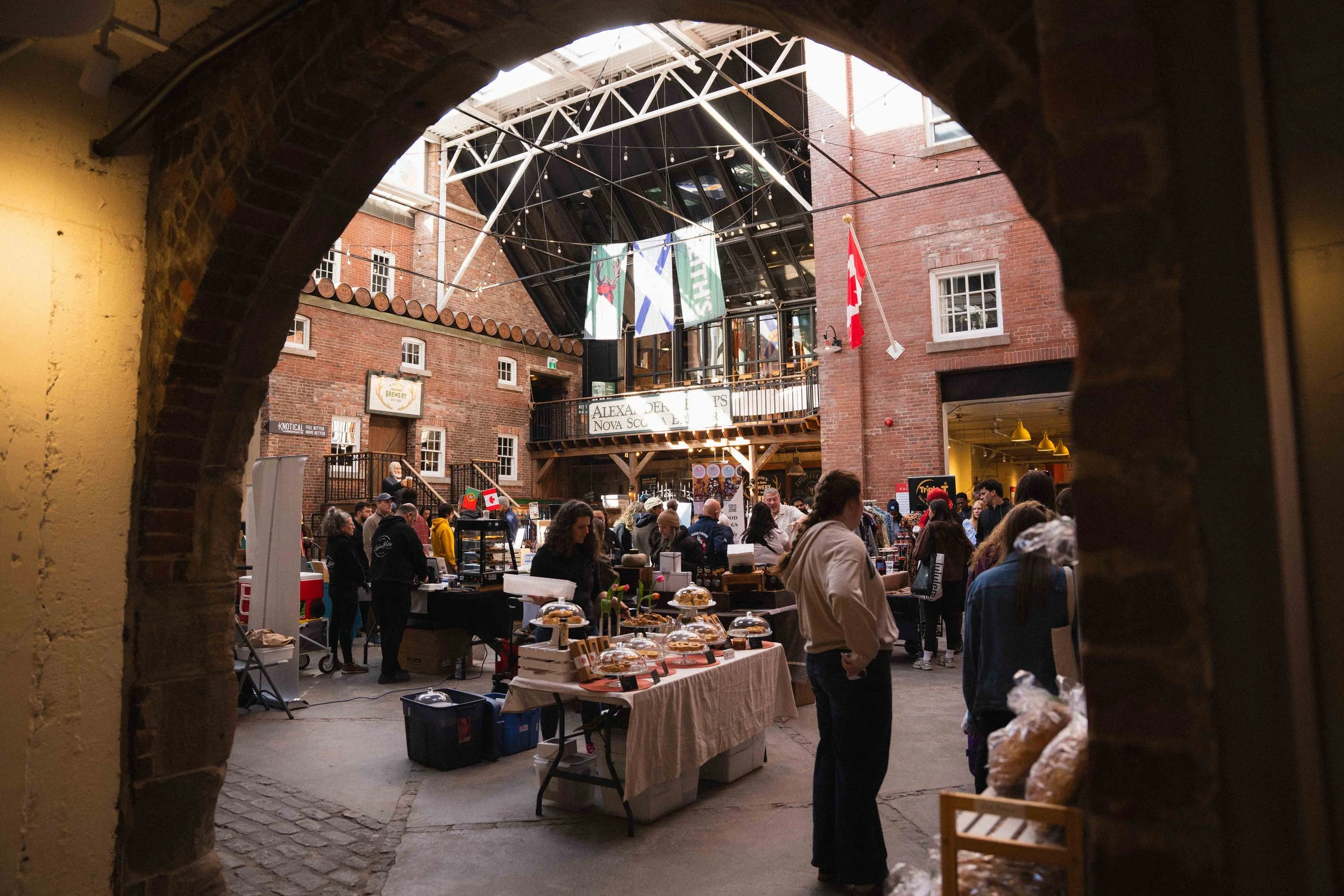 looking through an archway at a busy farmers' market in the Historic Alexander Keiths building near the Waterfront in Halifax, Nova Scotia. A Canadian flag and fairy lights are hanging from the ceiling, and the walls are brick and the floor is stone.