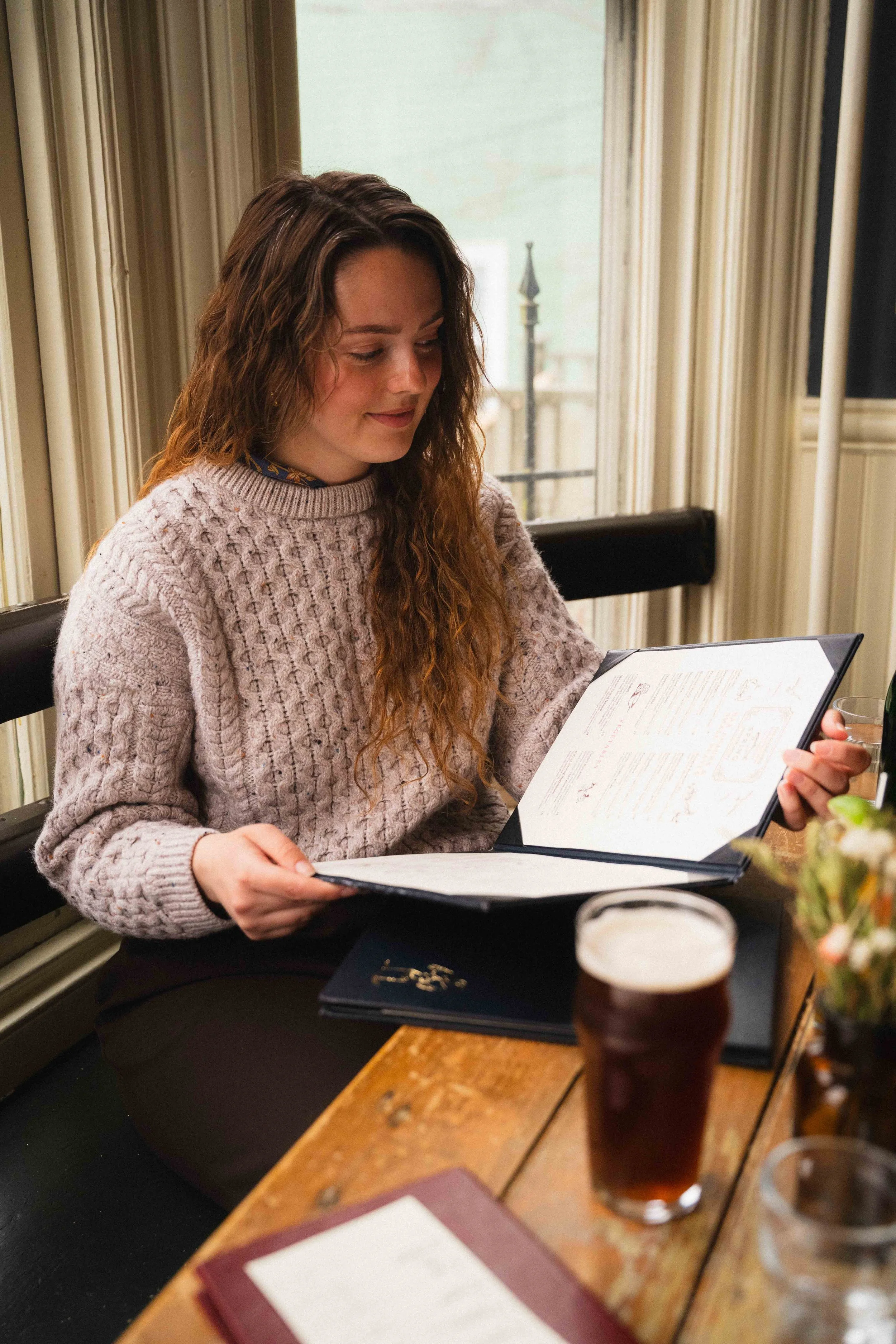 a woman reading a menu at a pub wearing a woollen knit sweater with a pint of beer in front of her as she is sitting in a window booth, restaurants in the city of Halifax, Nova Scotia