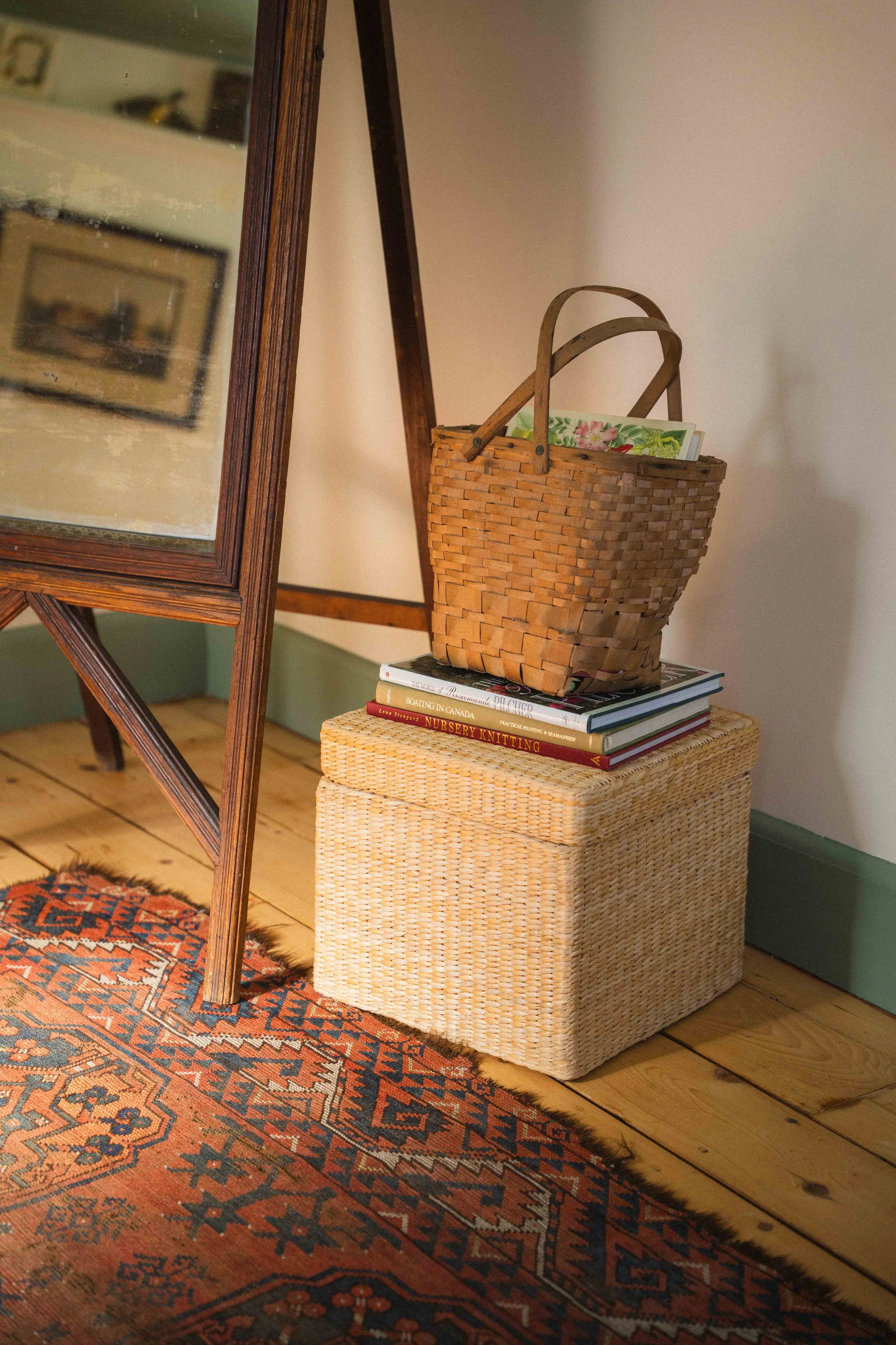 decorating an old home in the cottagecore aesthetic with colourful book stacks, including titles of Rosamunde Pilcher, Boating in Canada, and Nursery Knitting; paired with vintage woven baskets, wicker footstool, patterned wool rug antique mirror
