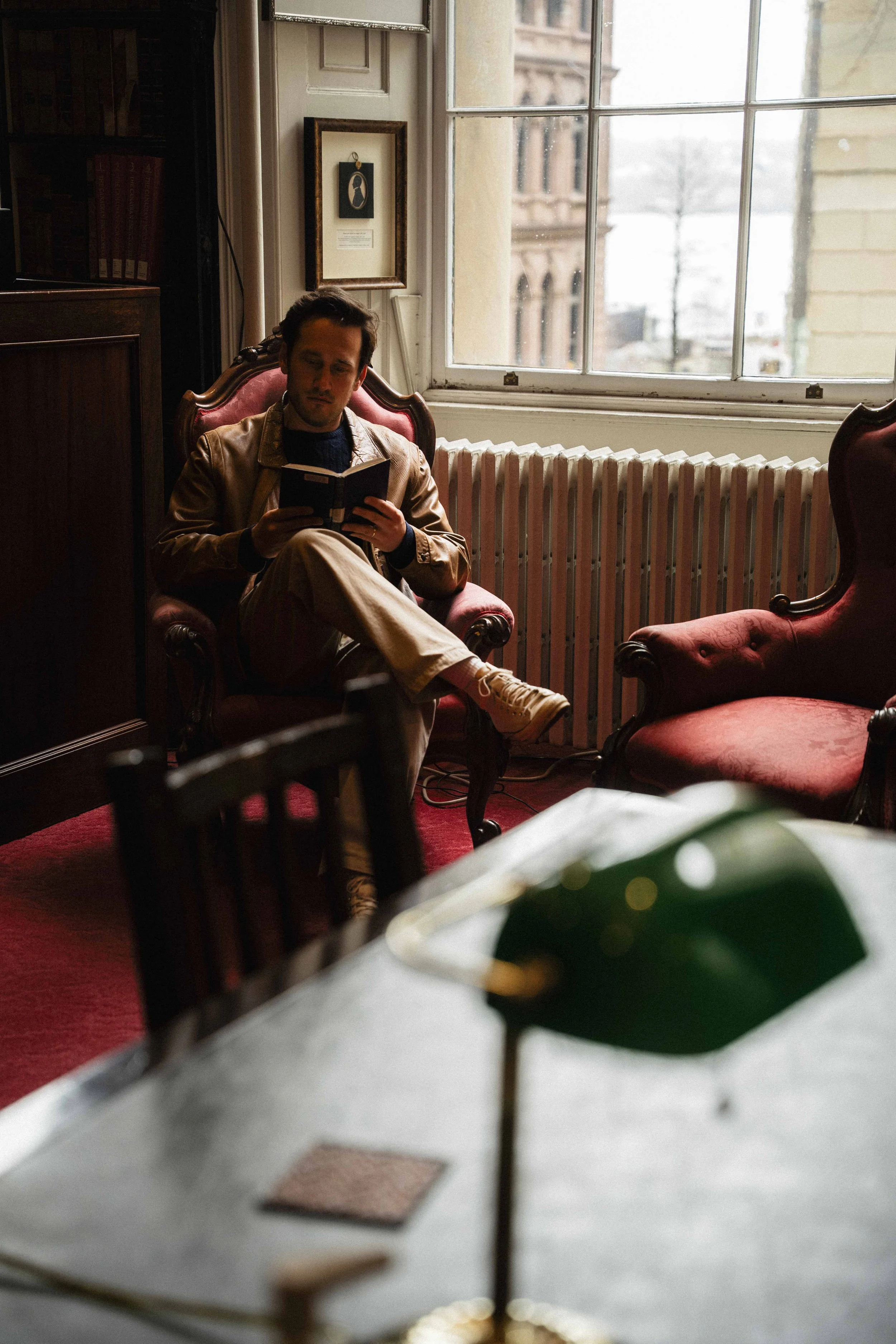 a man dressed in dark academia style reading an old book in an antique armchair  next to a large window and radiator with a vintage desk lamp in the foreground at a public library in Halifax, Canada