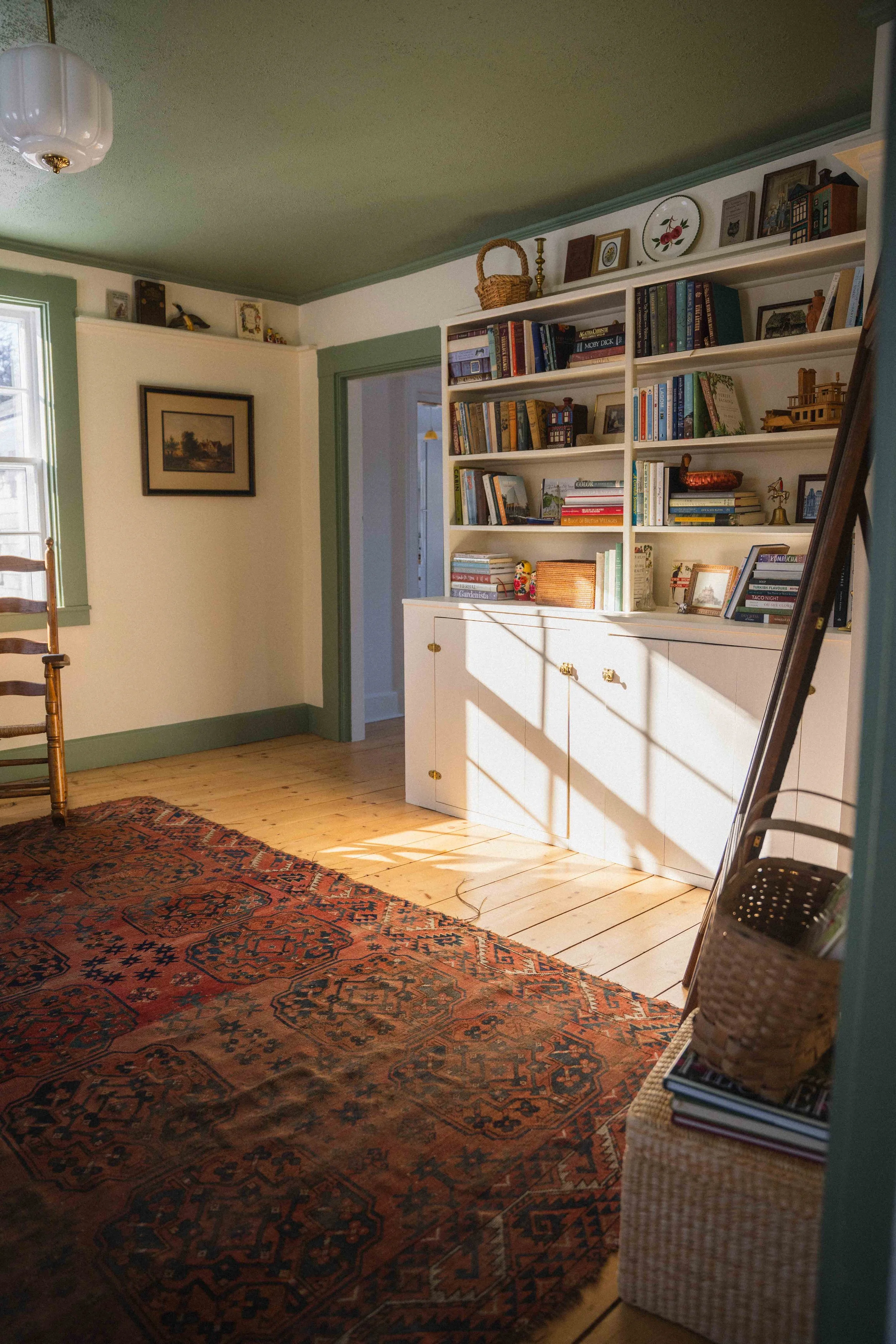 a cozy cottage library with cream walls, green ceiling and trim, an antique red wool turkish rug, a ladder back rocking chair, baskets, a colourful bookshelf, and oil painting