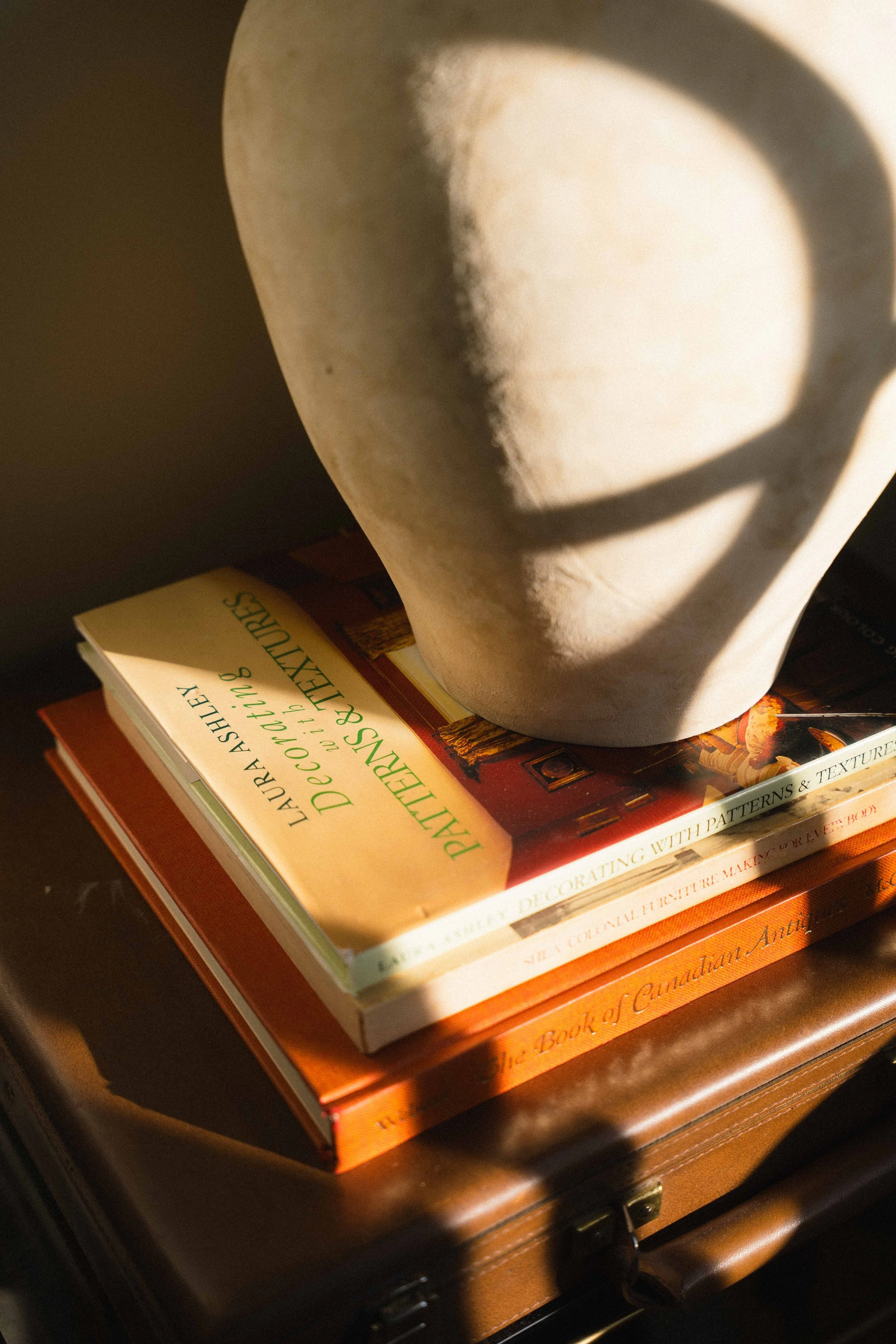film photo of sunlight streaming onto a stone vase with vintage book stack, including Laura Ashley's Decorating with Patterns & Textiles, Book of Colonial Furniture, and Book of Canadian Antiques for cottagecore home decor