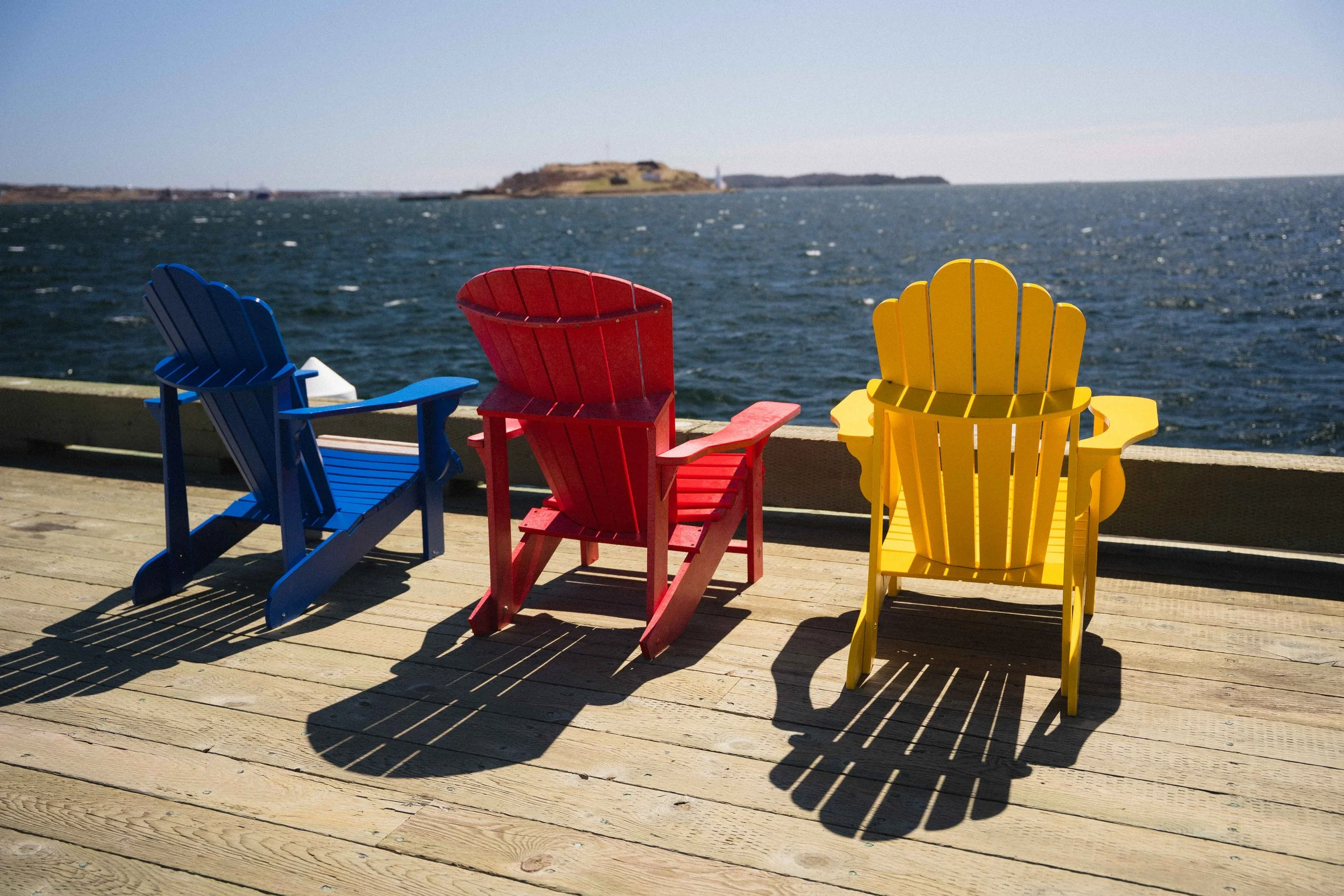 adirondack chairs in primary colours red, yellow, and blue looking out at the harbour on Halifax Waterfront boardwalk with lighthouse in the background by the Atlantic ocean for a guide to Third Spaces
