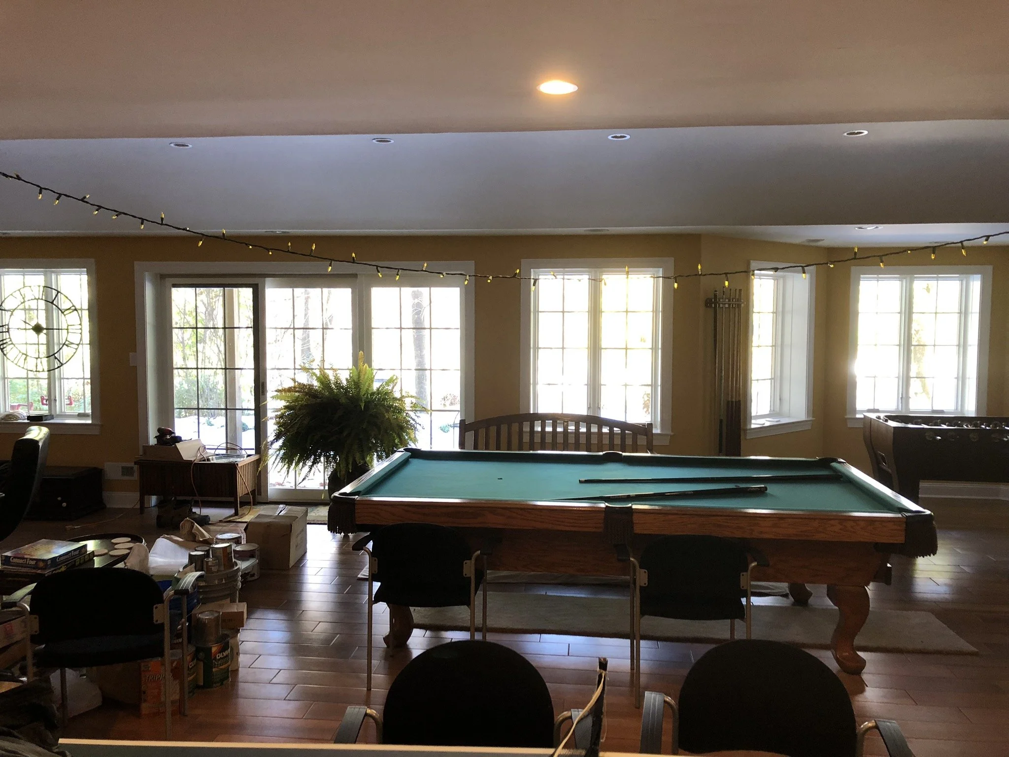 A game room with a pool table, foosball table, and a foosball table, lit by natural sunlight from multiple windows and decorated with string lights.