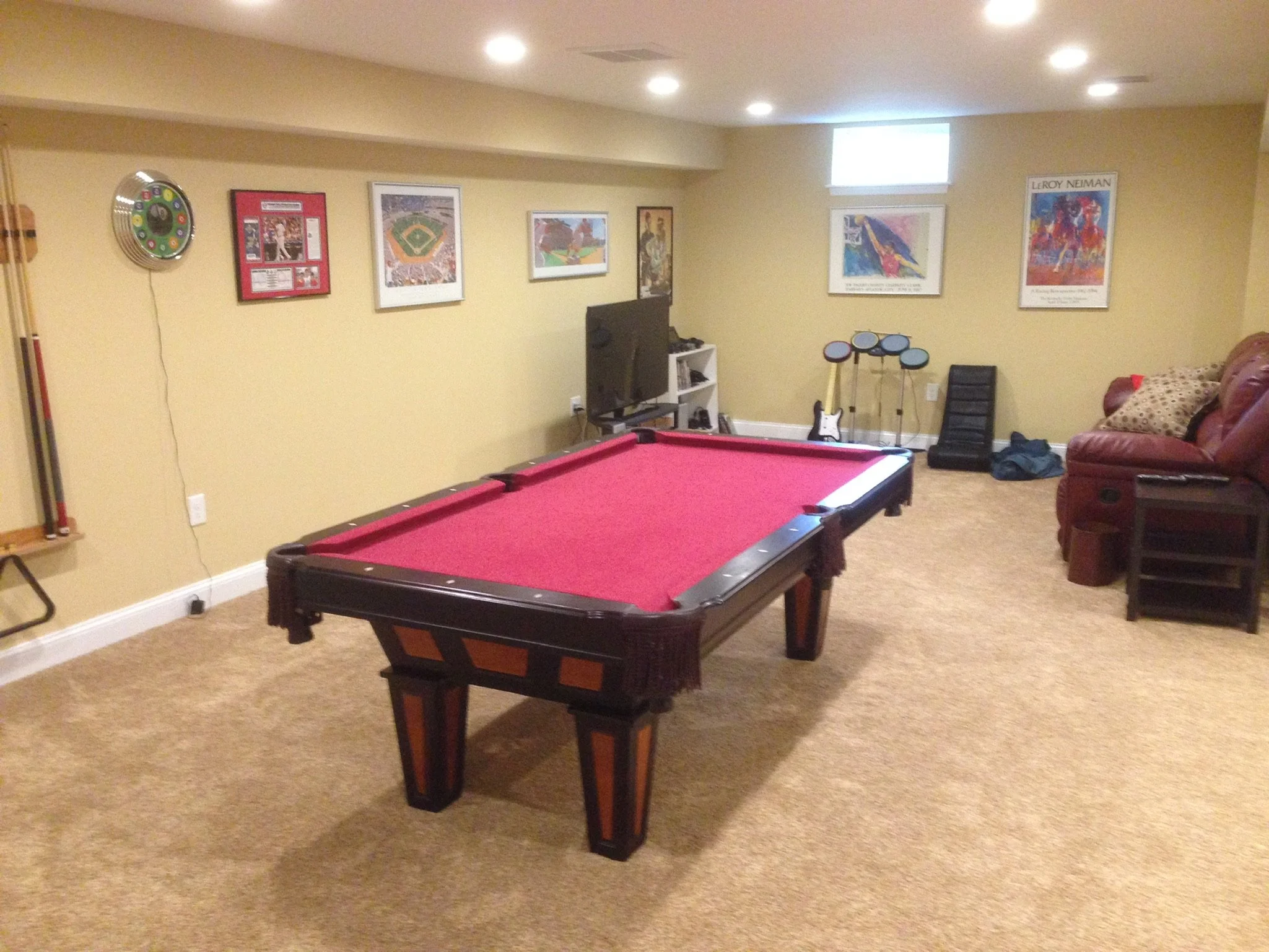 Basement game room with a pool table in the center, a wall-mounted dartboard, paintings, a television, a guitar, a video game controller, a small stereo, an armchair, and a couch.