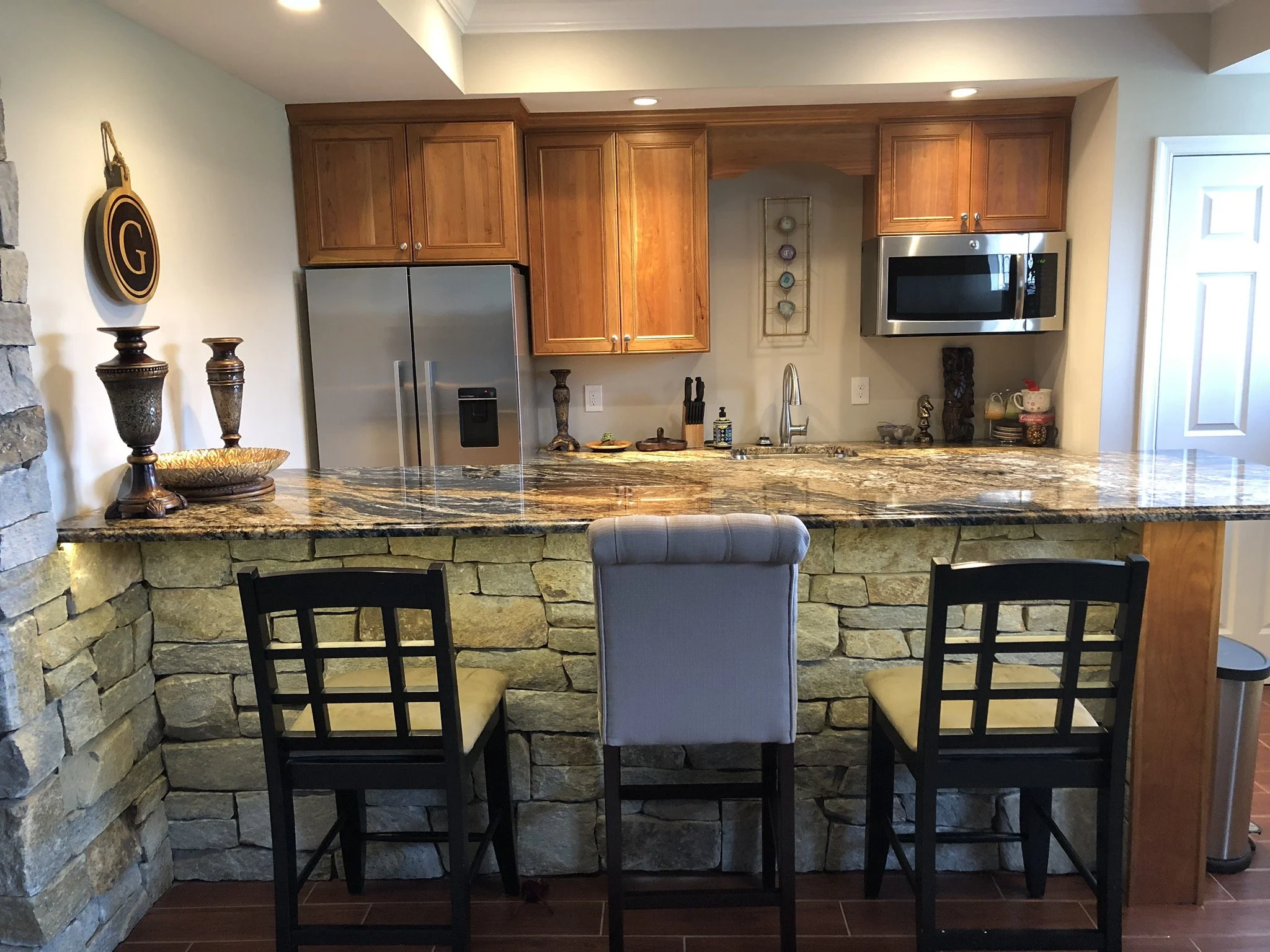 Kitchen with wooden cabinets, granite countertops, stainless steel refrigerator and microwave, stone-covered bar with three chairs, and various decorative items.