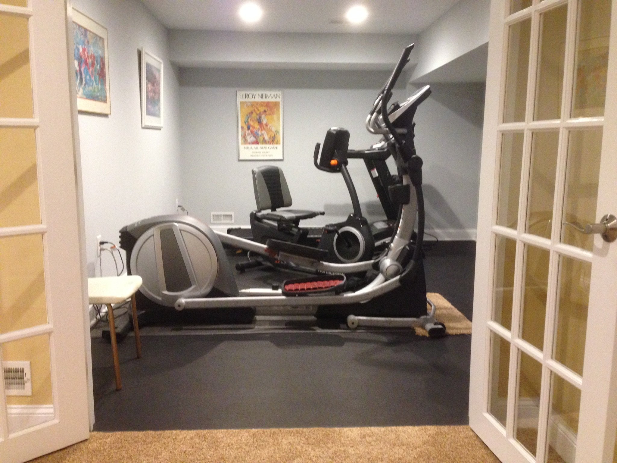 A home gym with a treadmill, an elliptical machine, and a chair, seen through glass-paneled double doors, with framed artwork on the walls.