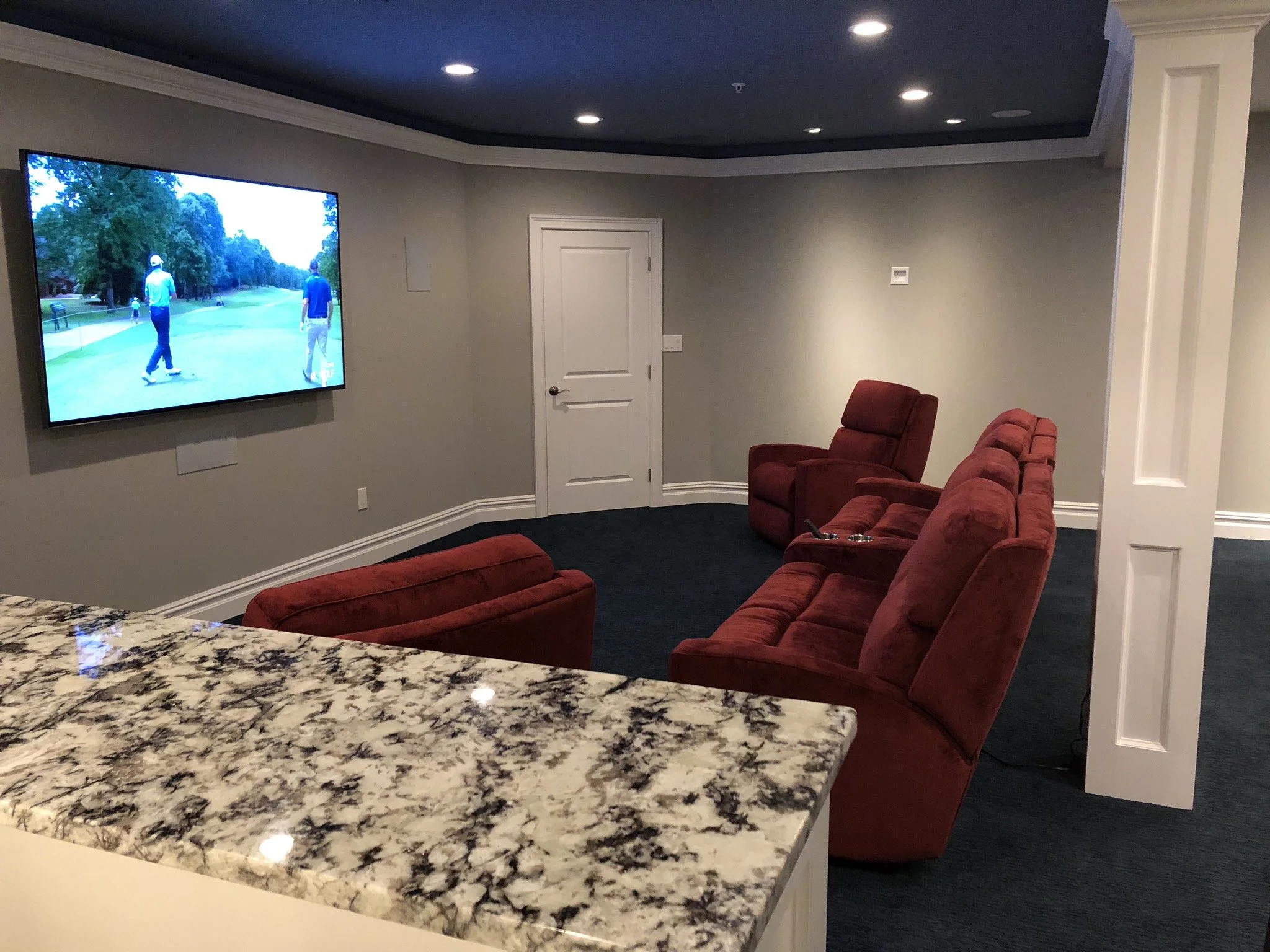 A cozy home theater with a large flat-screen TV showing a golf game, red recliner chairs facing the TV, a granite countertop in the foreground, and soft ceiling lighting.