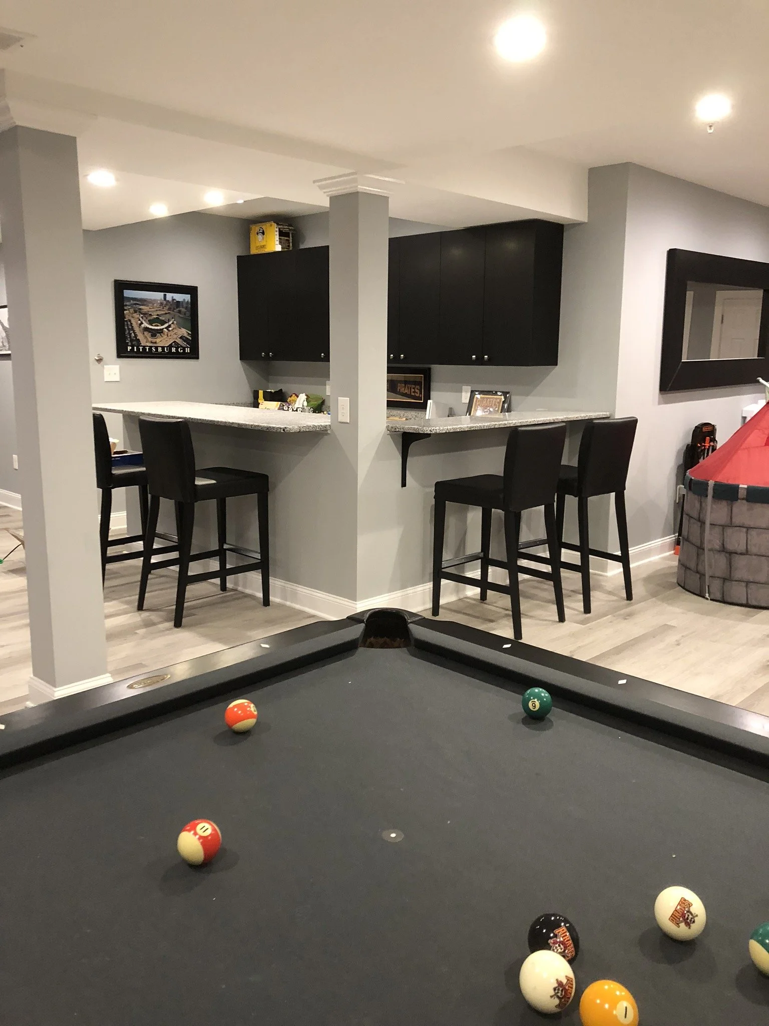 Indoor recreation room with pool table, balls on the table, bar with high chairs, black cabinets, and Pittsburgh-themed decor.