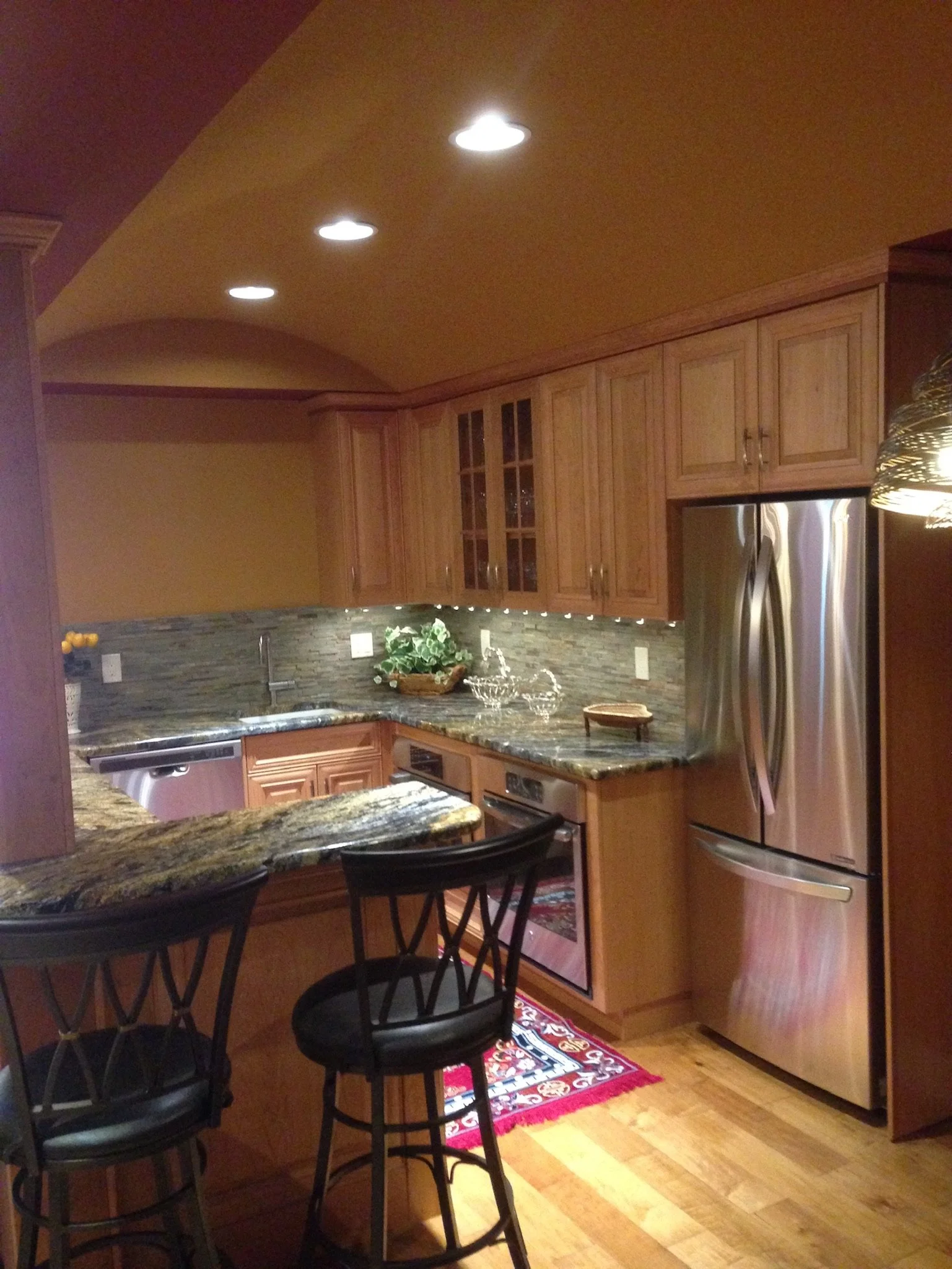 A kitchen with wooden cabinets, granite countertops, stainless steel appliances, a small floral rug, and black bar stools.
