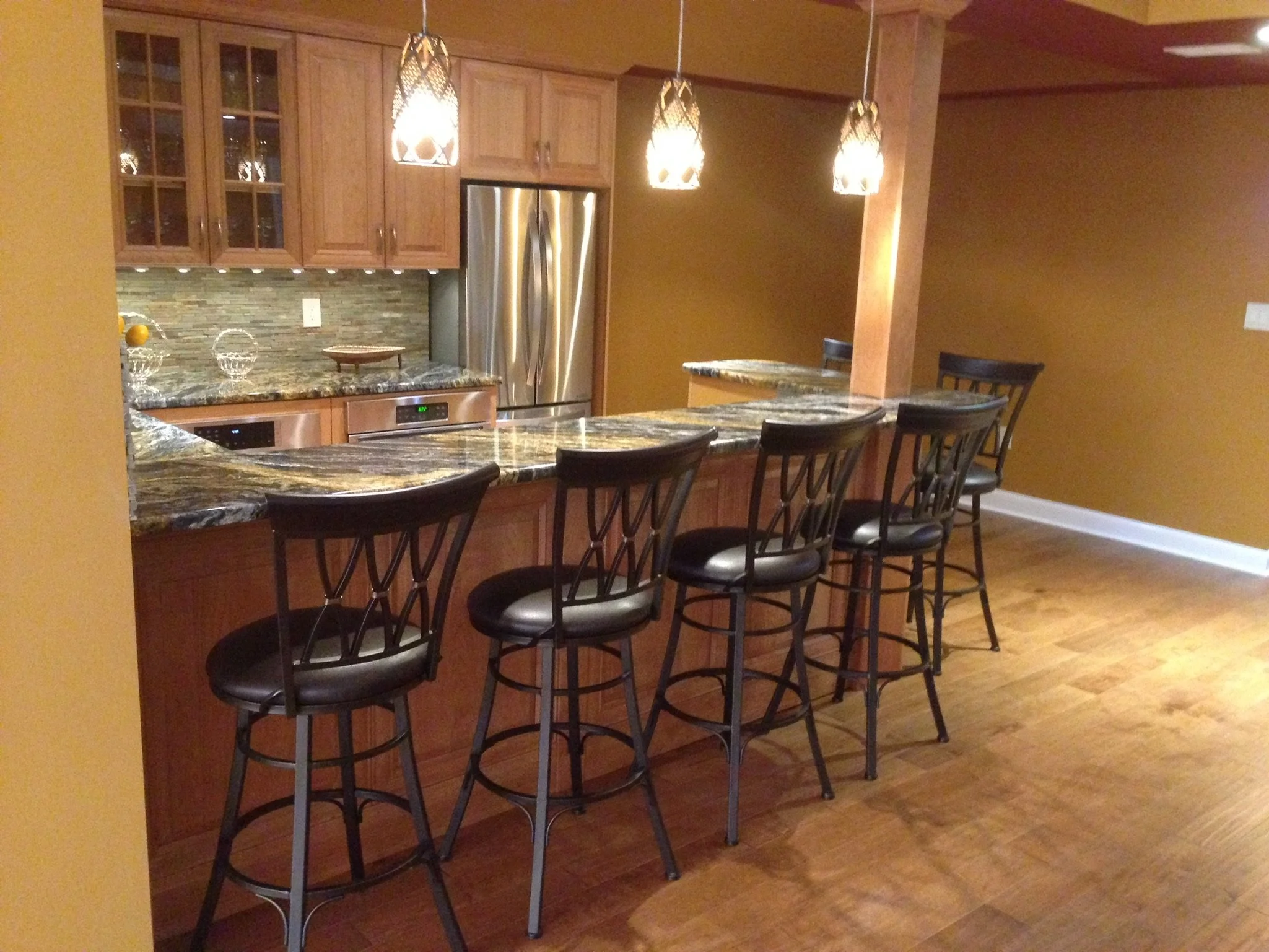 Kitchen with a granite countertop bar and five black barstools, wooden cabinets, a stainless steel refrigerator, and three hanging pendant lights.