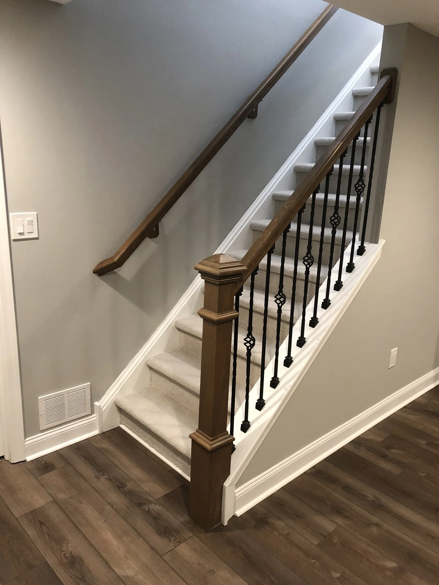 Interior staircase with beige carpeted steps, wooden handrails, black decorative metal balusters, hardwood flooring at the bottom, and a gray wall.