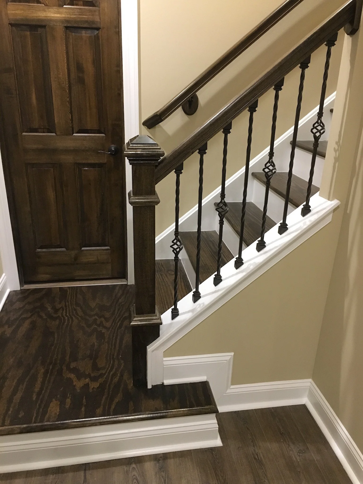Interior view showing a stair railing with dark wood handrail and decorative black metal spindles, a wooden door, and dark hardwood flooring in a house.