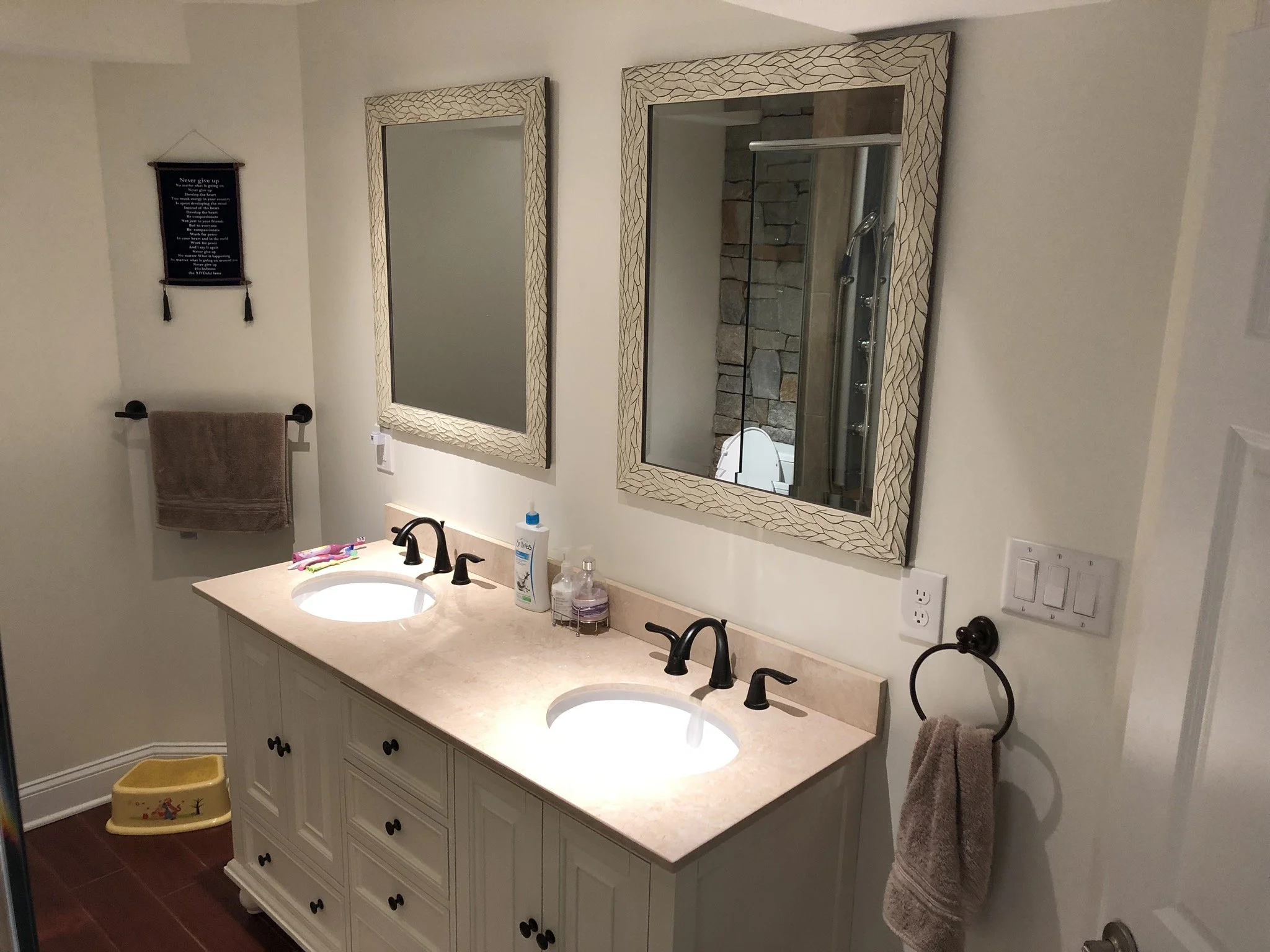A bathroom with a dual-sink vanity, two mirrors, hand towels, and children's toothbrushes and toys. The reflection in the right mirror shows a shower with stone wall and glass door.
