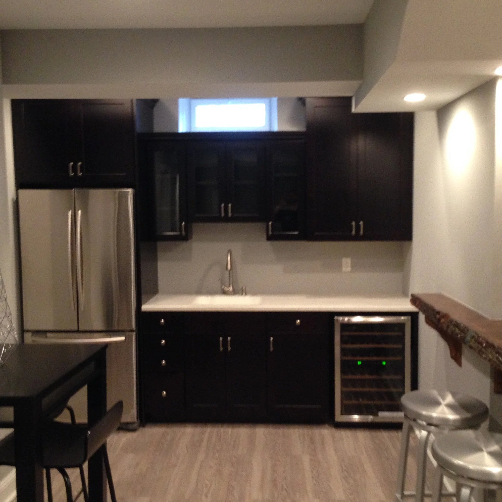 Modern kitchen with black cabinets, stainless steel refrigerator and wine cooler, white countertop, small window above sink, and overhead lighting.