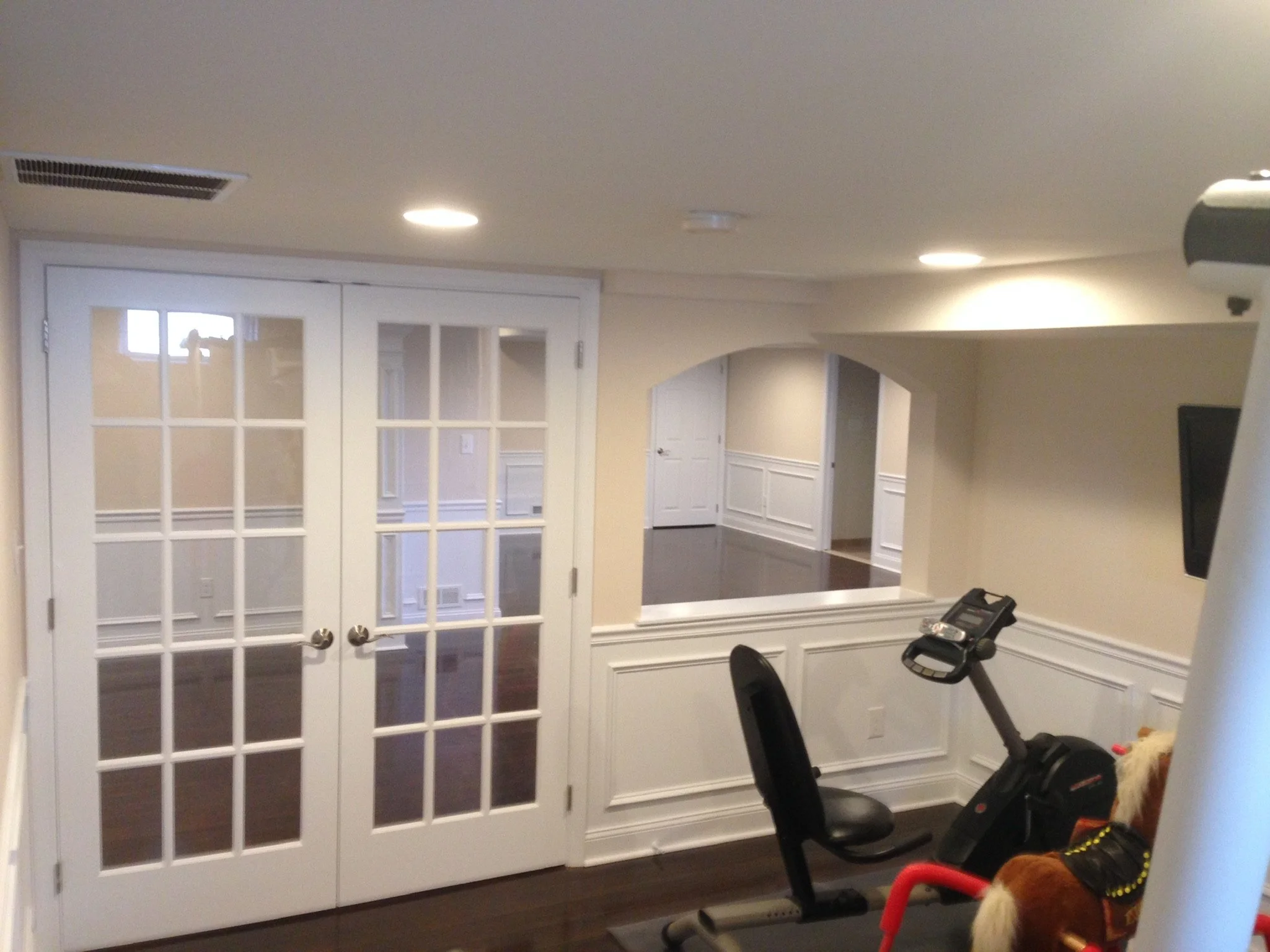 Room with glass double doors, an exercise bike, and a small rocking horse near the exercise equipment. The walls are cream-colored with white wainscoting, and there are ceiling lights.