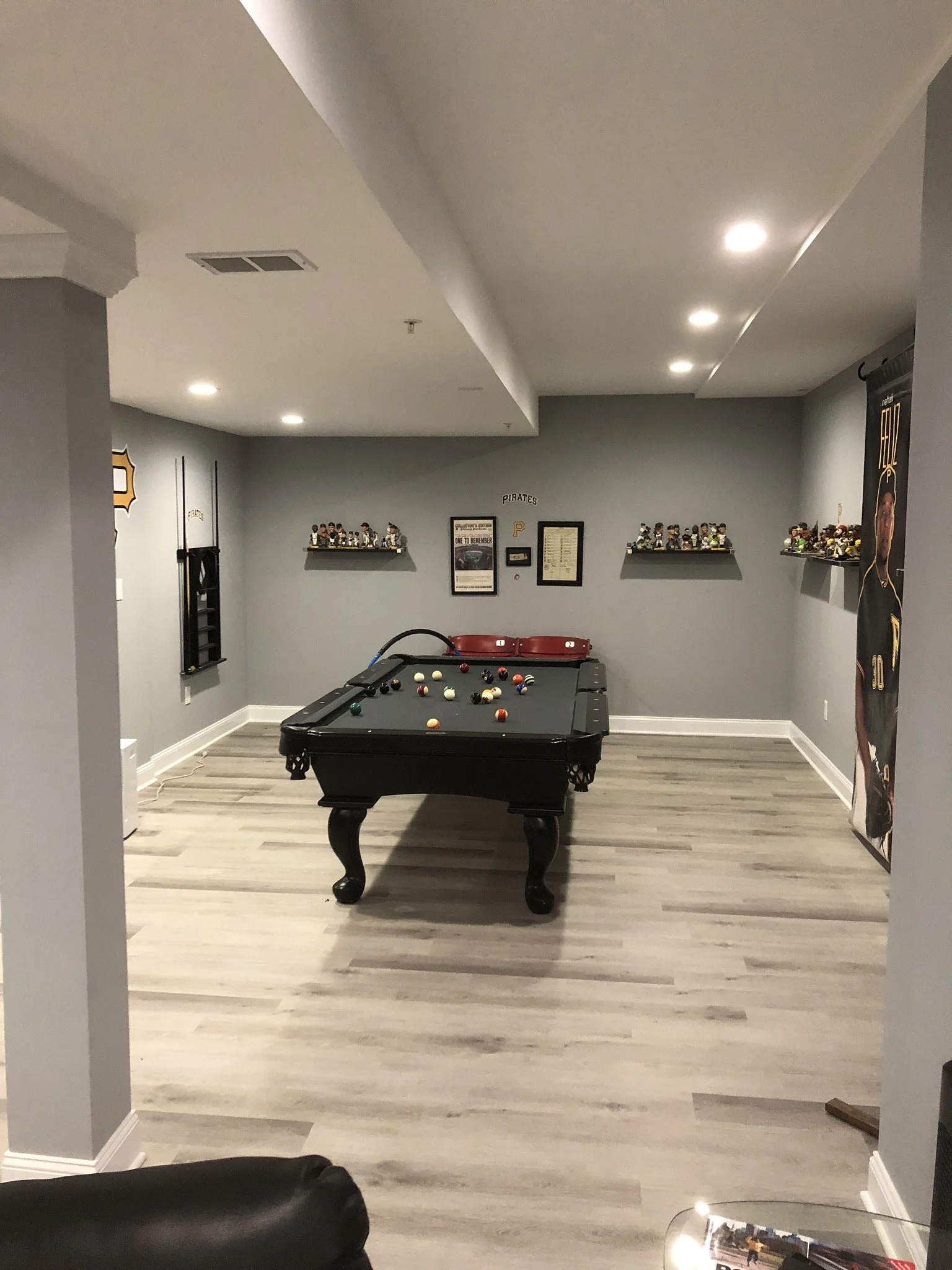 A basement entertainment room with a black pool table, framed pictures on the wall, shelves with figurines, and a Pittsburgh Pirates themed decoration.