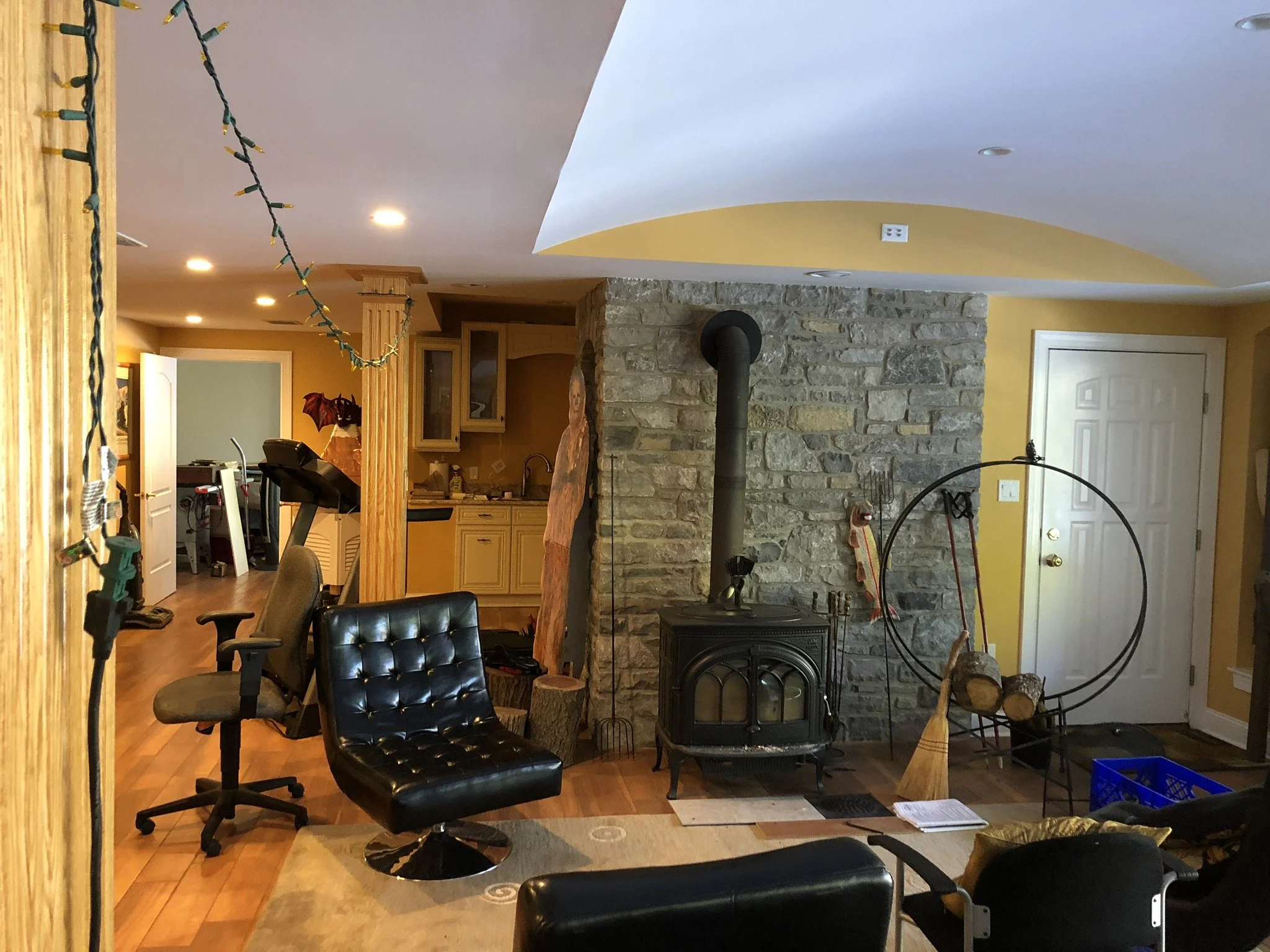 Living room with a wood stove in front of a stone wall, a black chair, and a large circle metal firewood holder. There are logs inside the holder and logs on the floor. An empty blue crate is near the wall, and a broom rests against the wall. The roo