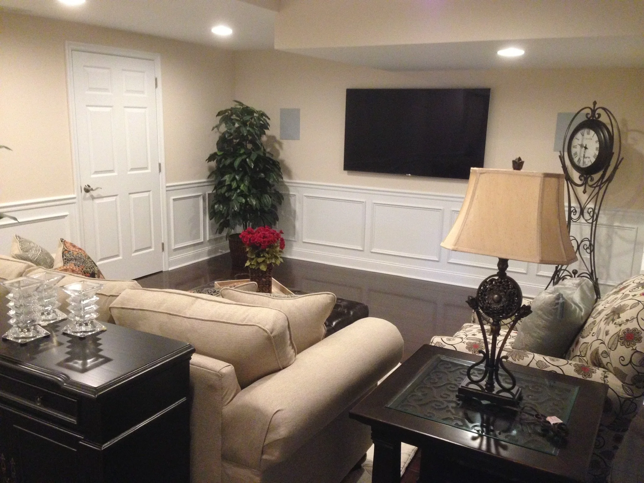 Living room with beige couch, patterned armchair, dark wood table, potted plant, wall clock, flat-screen TV, table lamp, and decorative pillows.