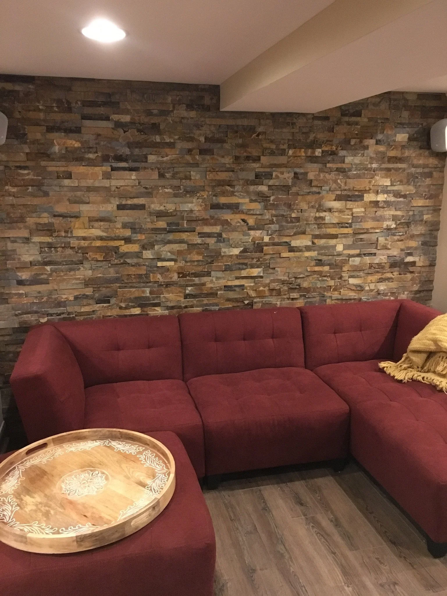 A living room corner with a red sectional sofa, a round wooden tray on the armrest, a yellow throw blanket, a brick accent wall, a ceiling light, and wooden flooring.