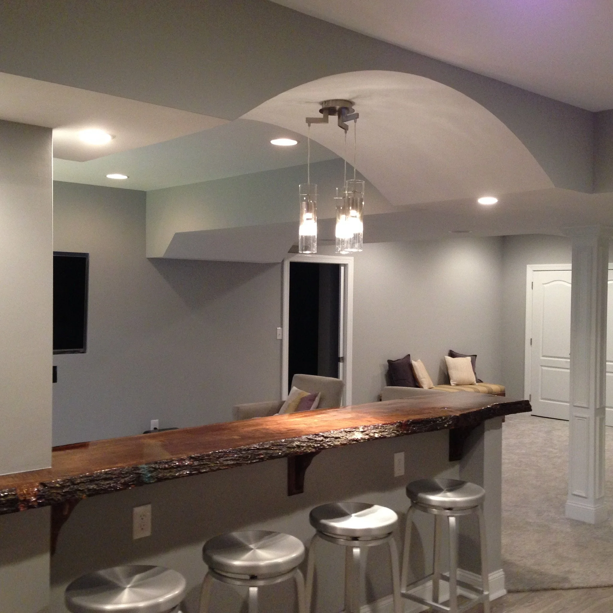 Interior of a basement with a wooden bar counter, four silver bar stools, a seating area with pillows, and ceiling lights.