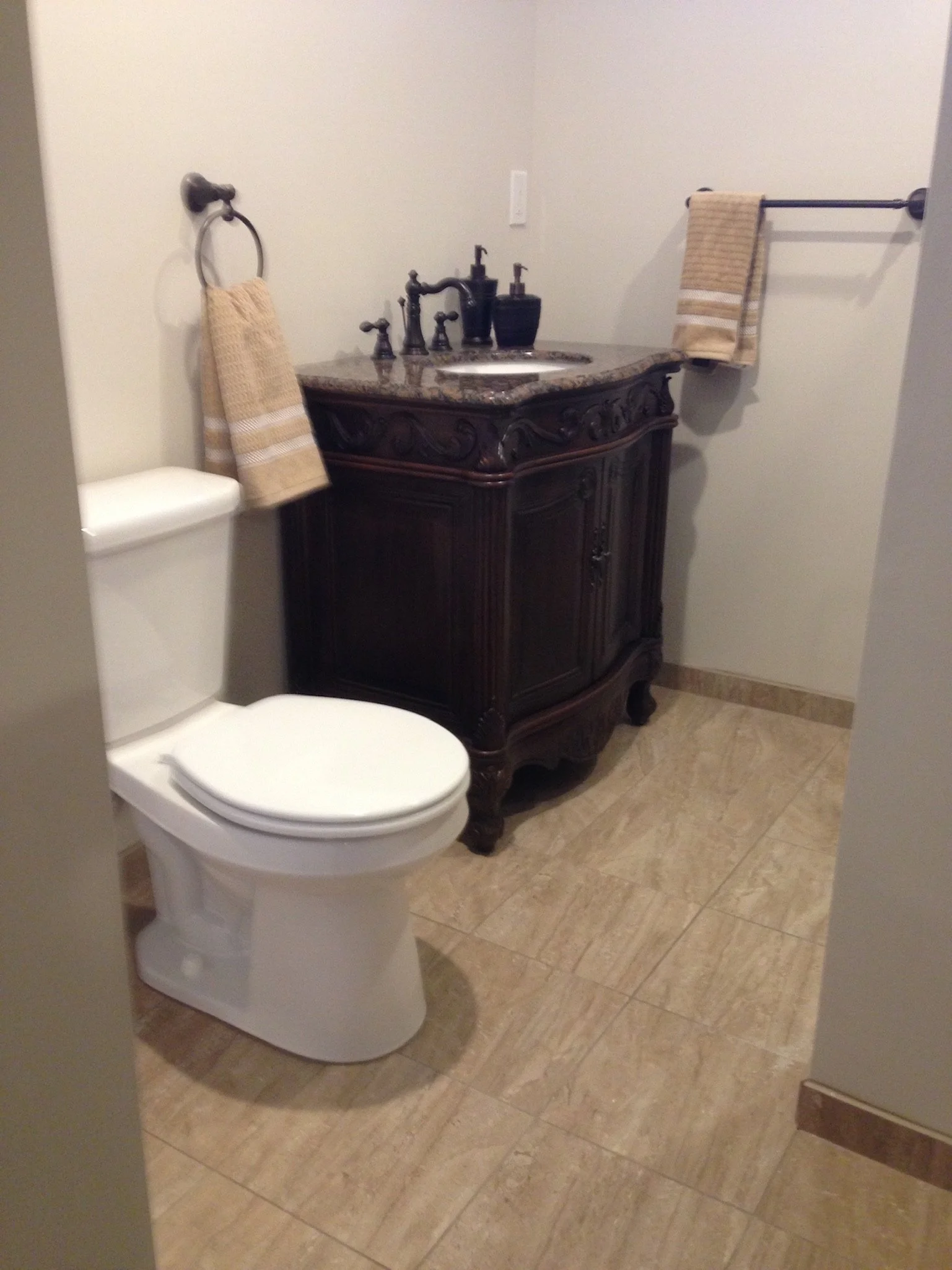A small bathroom with a toilet and a dark wood vanity with a granite countertop, two soap dispensers, and two beige towels hanging on towel rings and a towel bar.