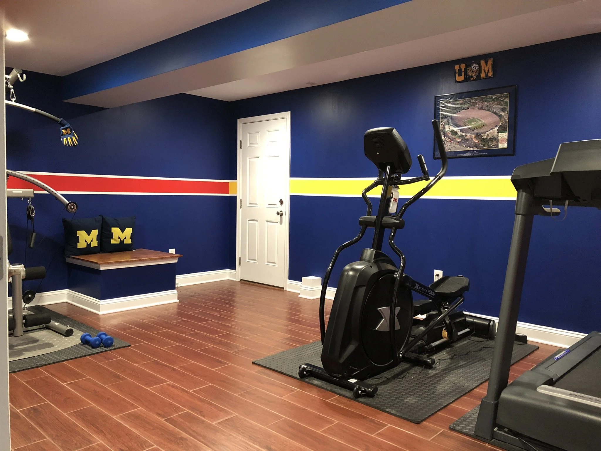 Home gym room with fitness equipment, including an elliptical machine, treadmill, and weights, decorated with Michigan Wolverines themed pillows and wall art.