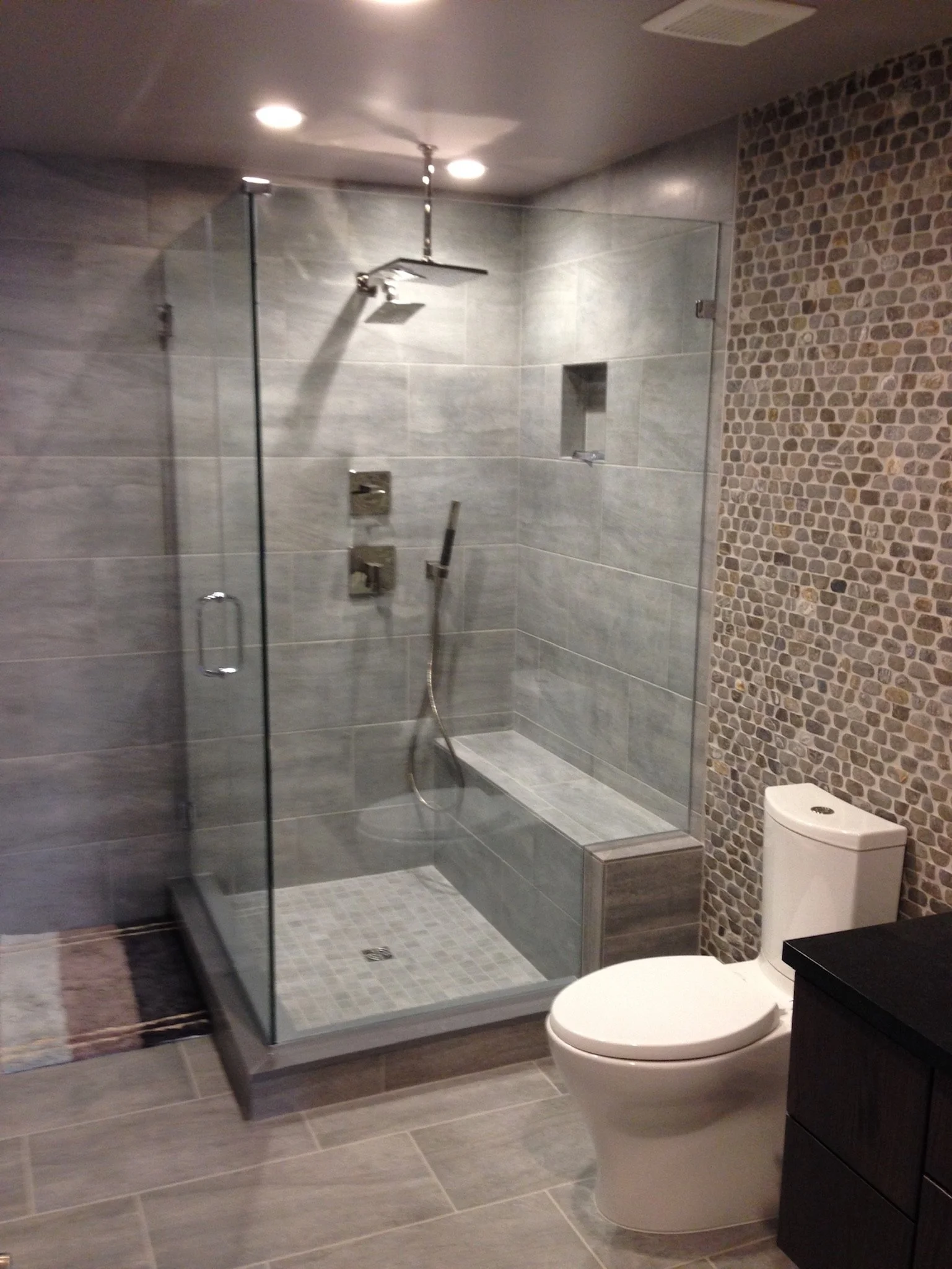 Modern bathroom with a glass shower enclosure, rainfall showerhead, and a toilet next to a wall with pebble tile design. The shower has gray tiles and built-in niches, with a small leaded mat on the tiled floor outside.