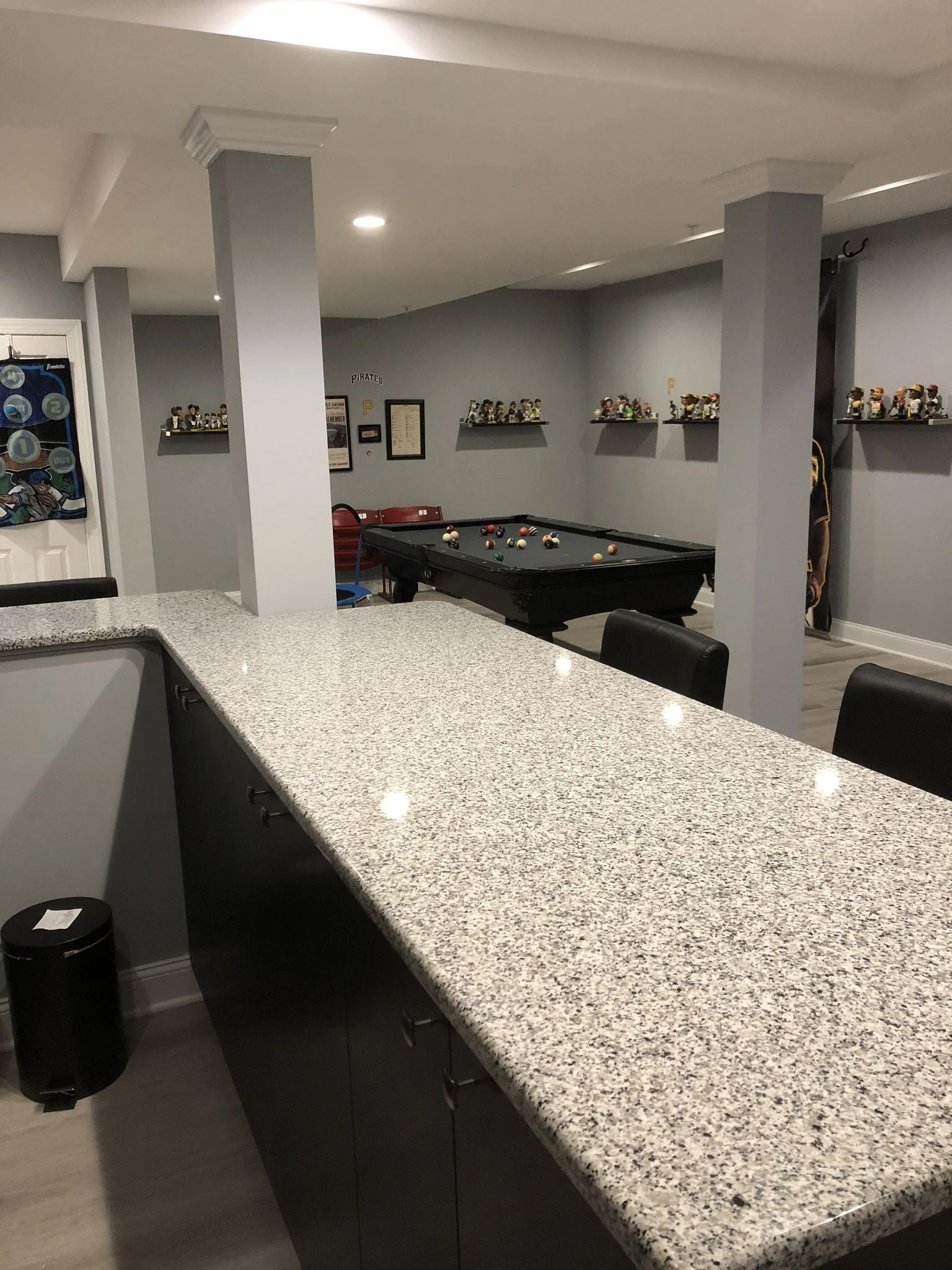 A recreational room with a pool table, a counter with a granite top, and shelves displaying bobblehead figures on the wall.