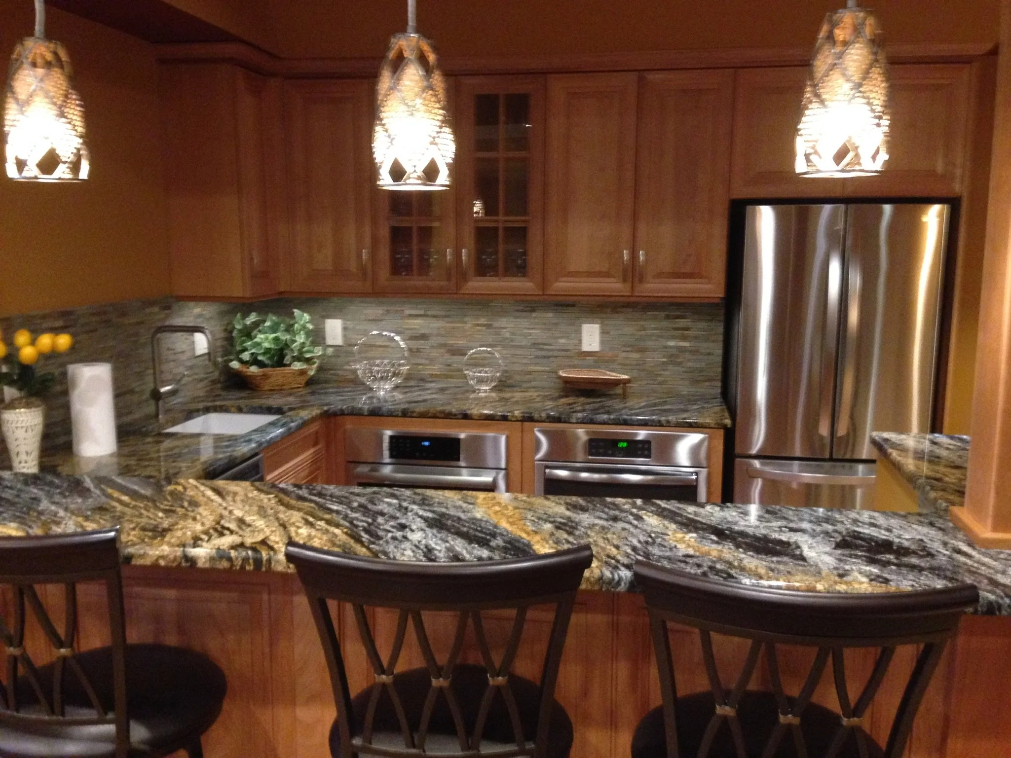 Kitchen with wooden cabinets, stainless steel refrigerator, granite countertops, pendant lights, and bar stools.