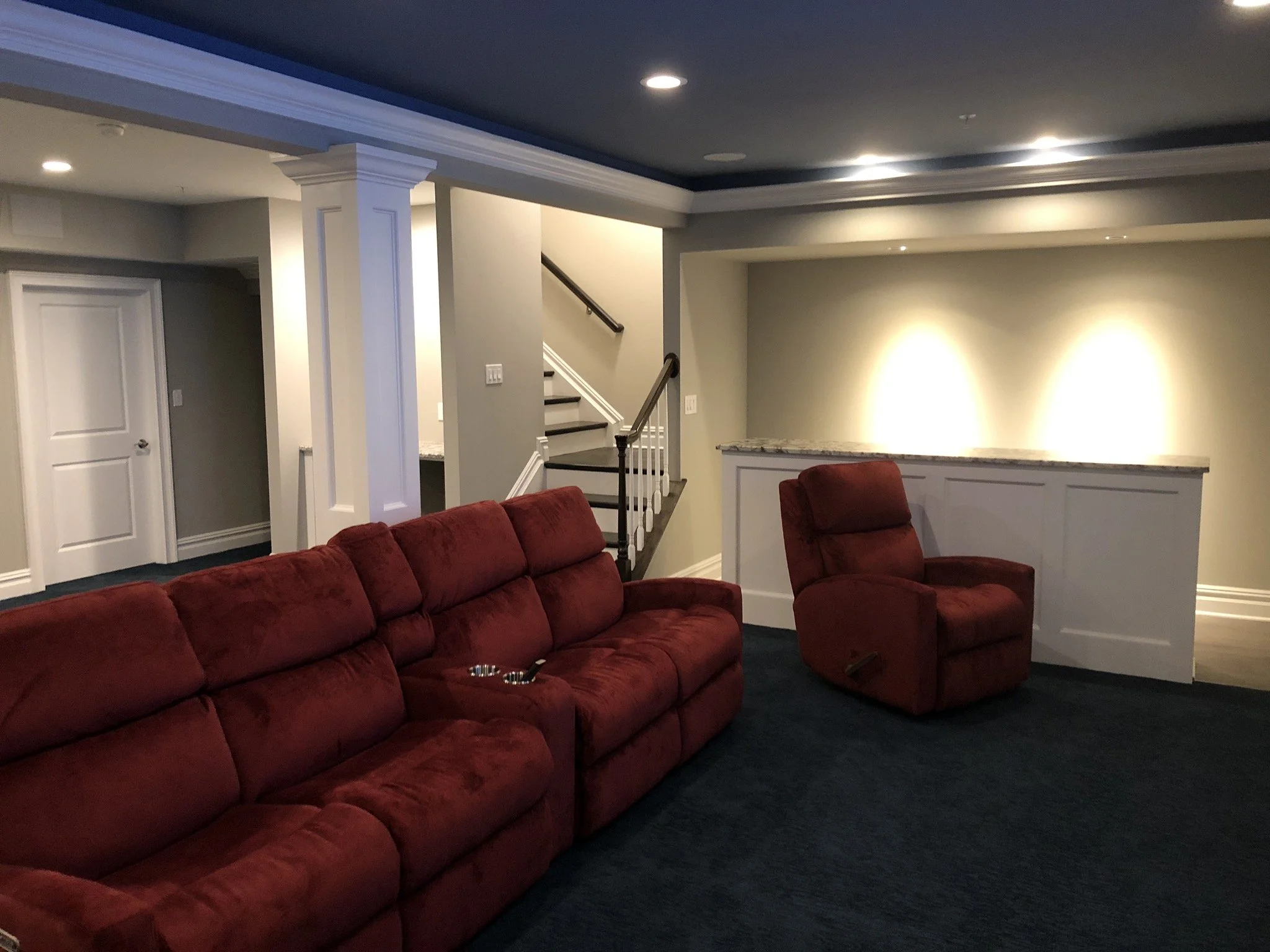 Living room with a red sofa and matching armchair, staircase in the background, recessed lighting, dark blue carpet, and light-colored walls.