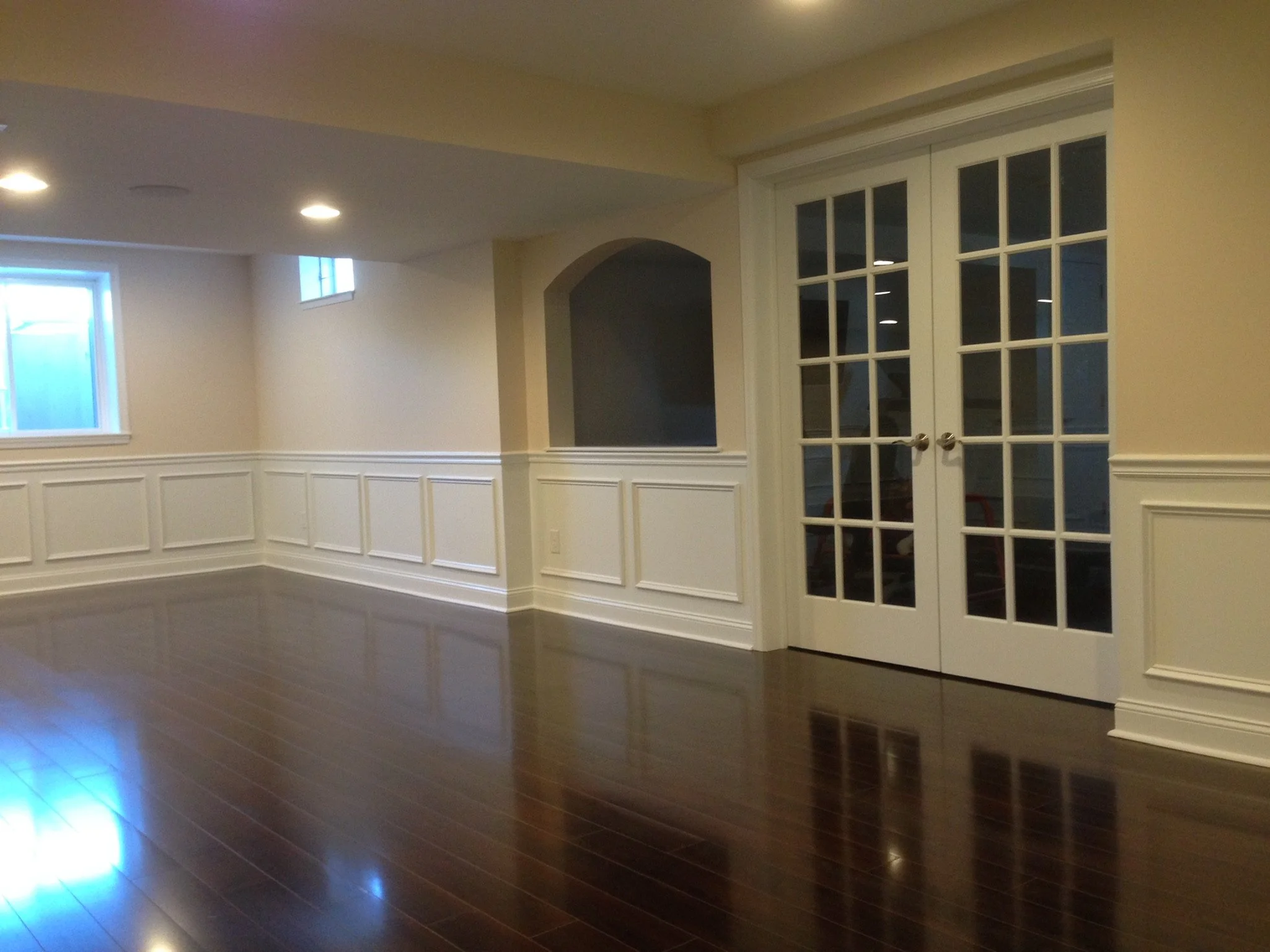 Empty living room with dark wood flooring, beige walls, white wainscoting, large windows, and closed glass double doors.