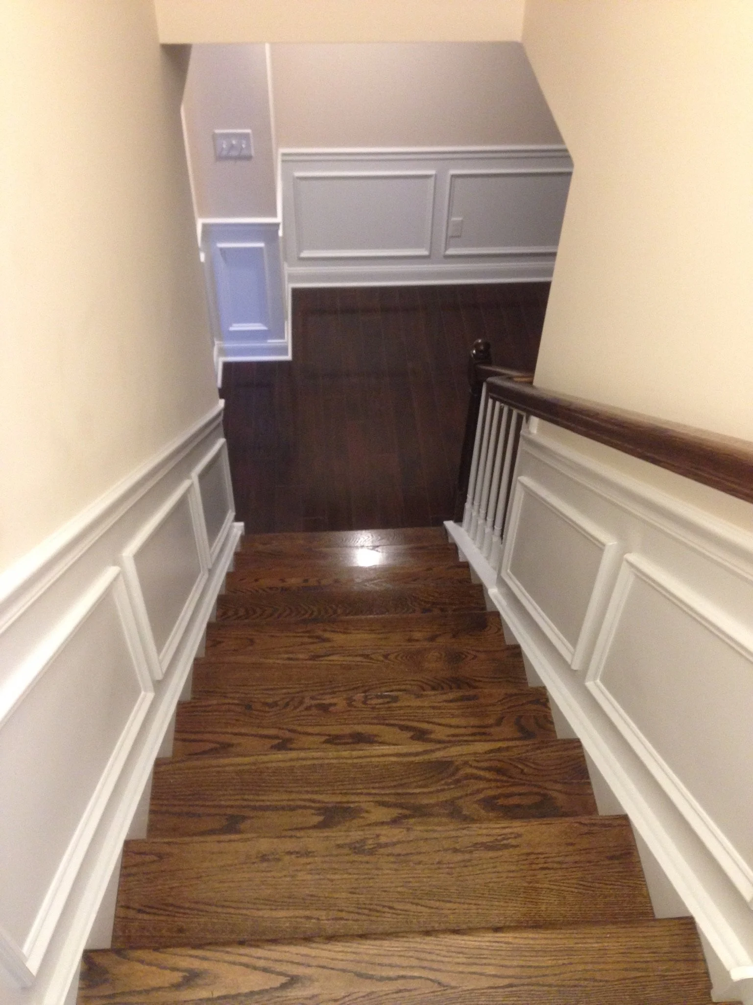 View of wooden staircase with white wainscoting, leading down to a room with dark hardwood flooring and white paneled walls.