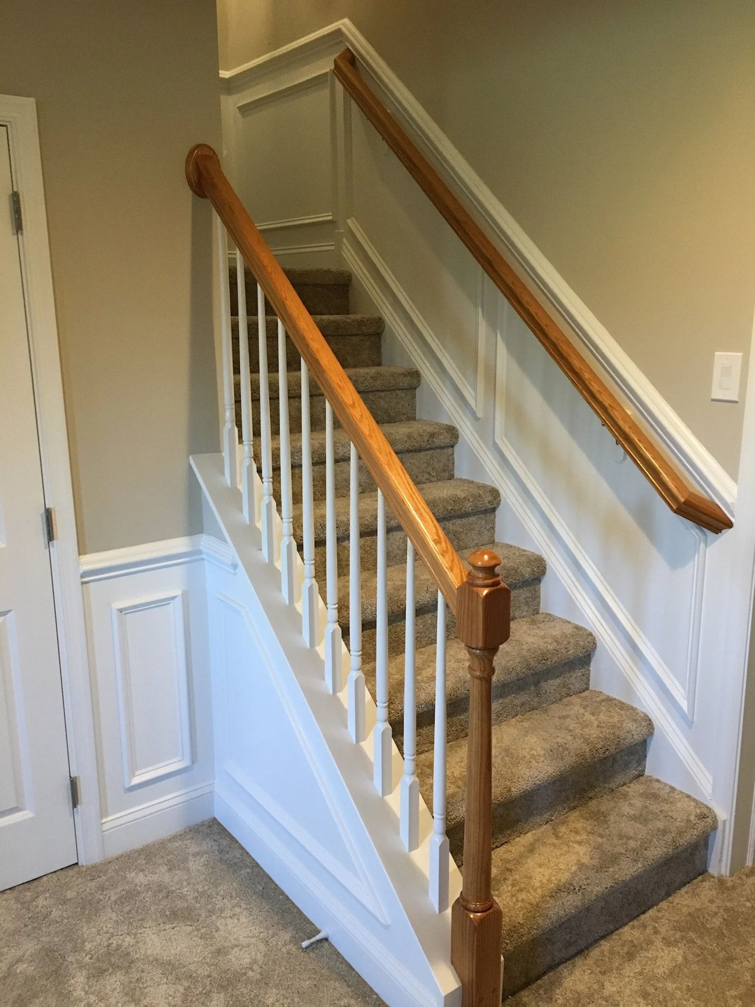 Interior staircase with beige carpet, white wooden railing with turned balusters, and a wooden handrail. The wall is painted light gray and has decorative molding. There is a closed white door on the left side.