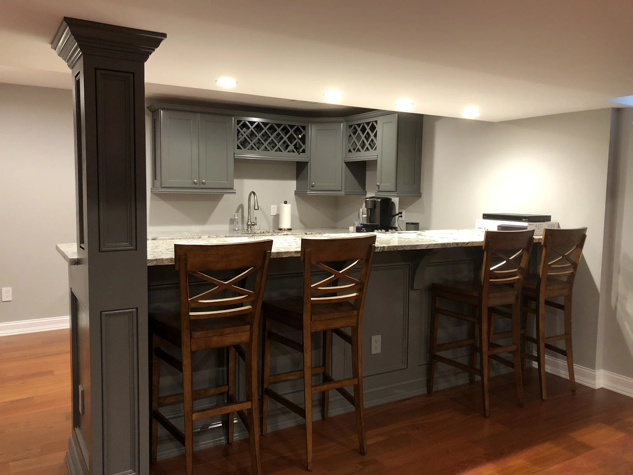 Gray kitchen bar area with four wooden bar stools and a granite countertop, with cabinetry, a sink, and small appliances in the background.