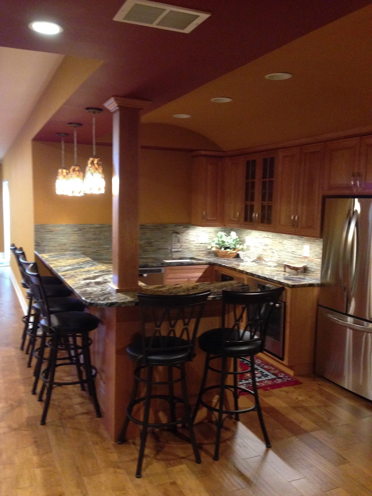 Kitchen with wooden cabinets, granite countertops, stainless steel refrigerator, and black bar stools around a counter island.