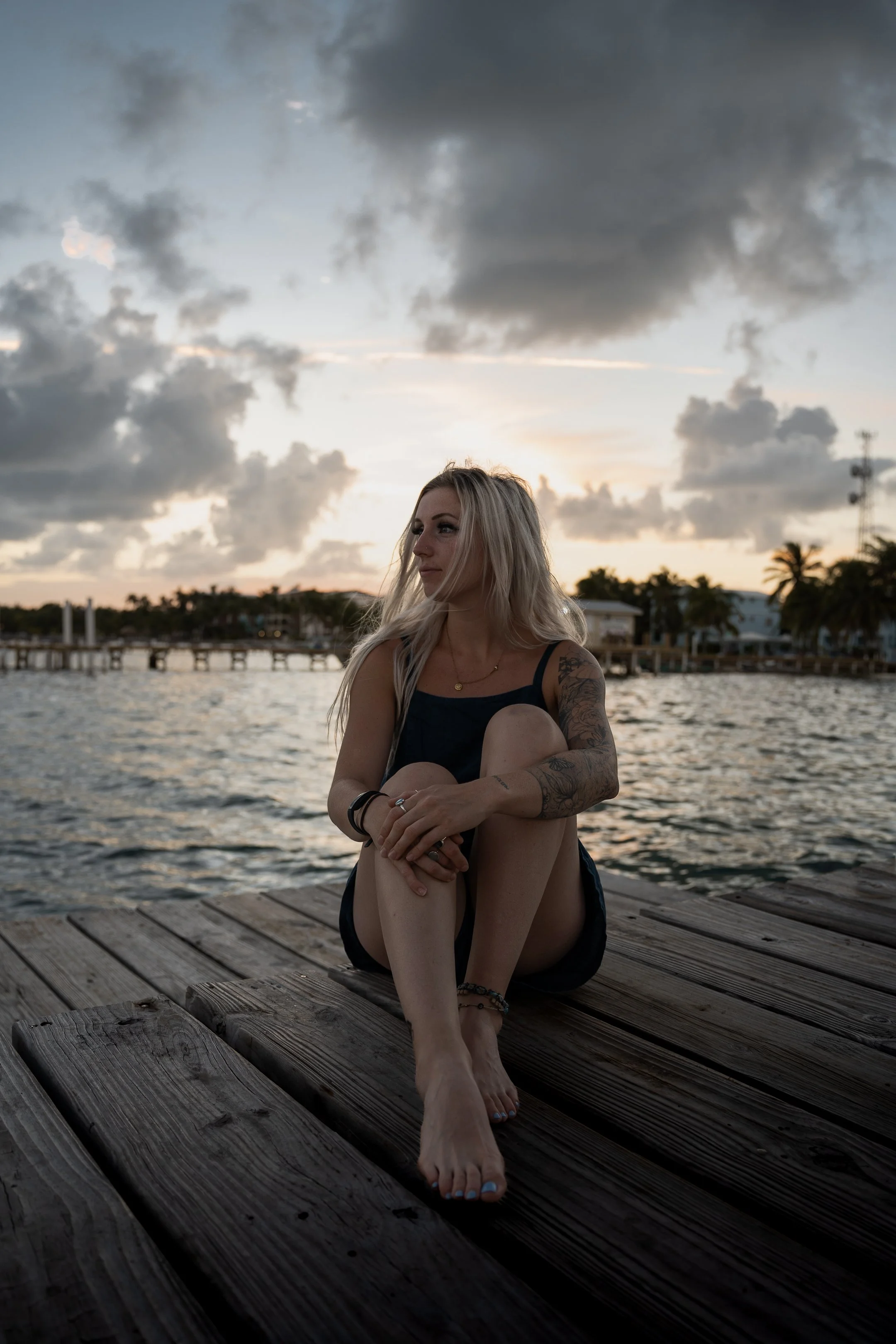 A young woman with blonde hair, sitting on a wooden dock near the water during sunset, wearing a dark dress or top, with tattoos on her arms, looking to the side.