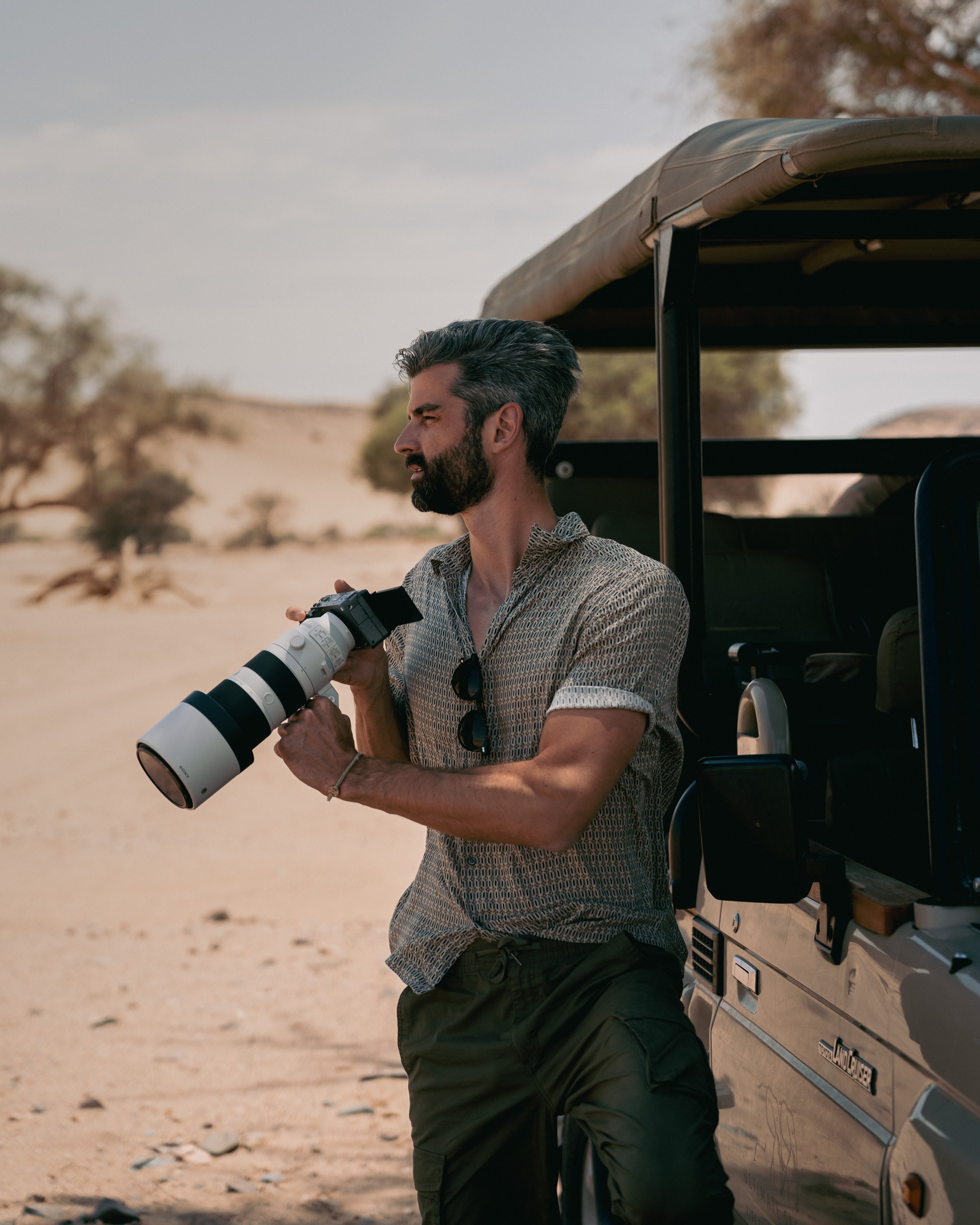 A man with a camera, standing next to a safari vehicle in a desert landscape with sparse trees during daytime.