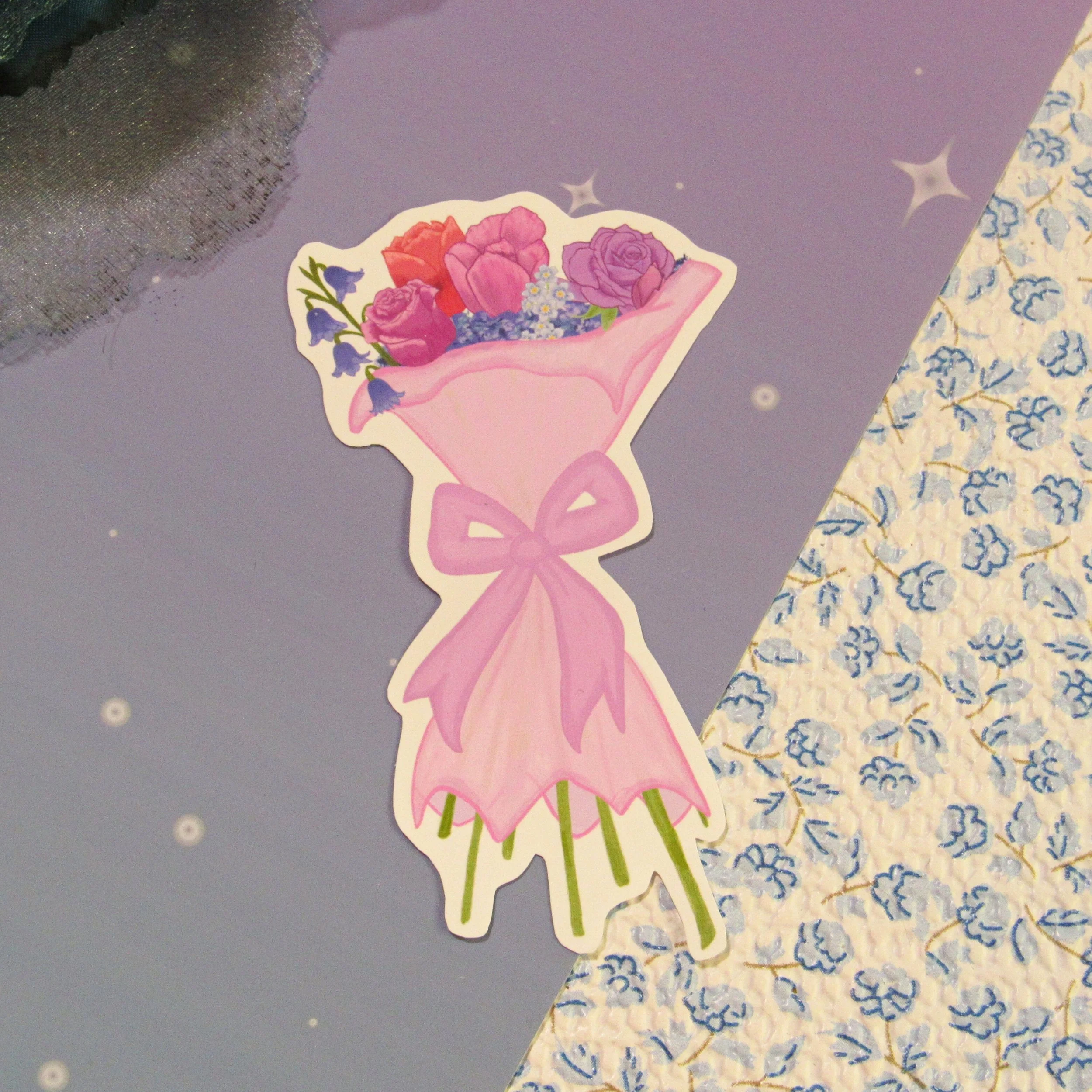 A flower bouquet sticker that has mostly roses in a bag, bluebell and blue flowers can also be seen in the sticker. The wrapping of the bouquet is pastel pink and wrapped in a large pink bow. At the bottom of the sticker pastel green stems.