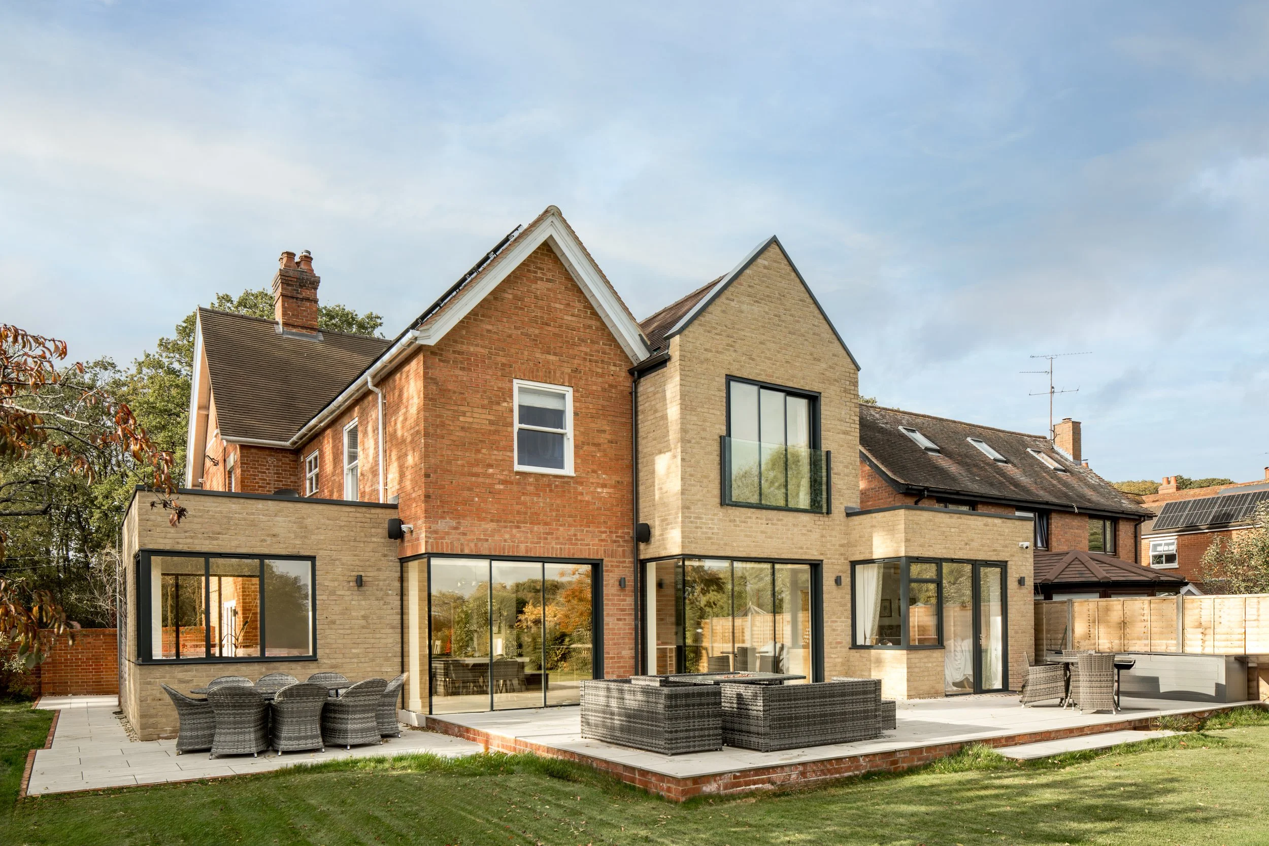 Pont + Wils Architects — Modern backyard patio with outdoor furniture, large glass windows, and a mix of brick and beige walls, with a lawn and neighboring houses in the backg